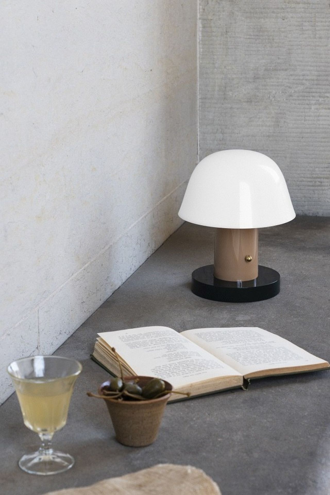AndTradition Setago Tischleuchte JH27, weiße Pilzlampe neben Buch, Getränk und Kapern auf grauem Untergrund.