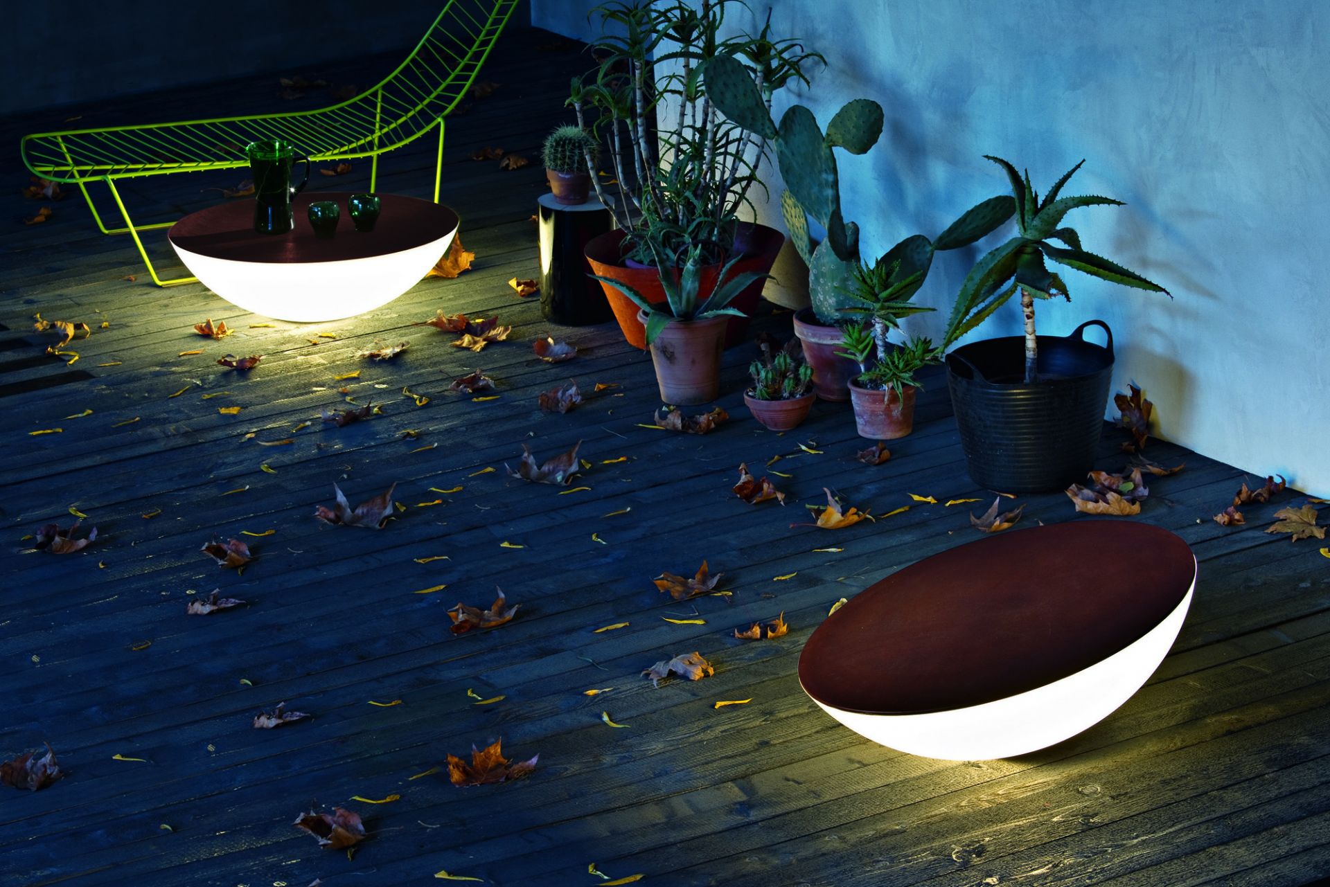 SOLAR Outdoor Bodenleuchte von Foscarini auf dunkler Holzterrasse mit Herbstlaub und Topfpflanzen.