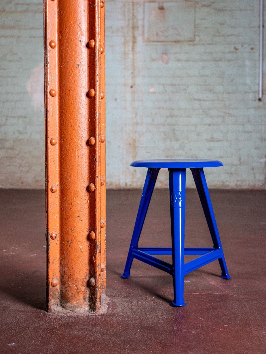 Blauer Rowac Hocker Modell IS 48 cm, ein robuster Industriehocker aus Metall neben einer Säule.