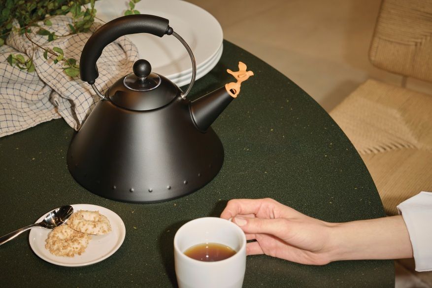 Schwarzer Alessi 9093 BB Wasserkessel auf Tisch mit Tasse Tee und Gebäck. Design-Wasserkocher mit Vogelpfeife.