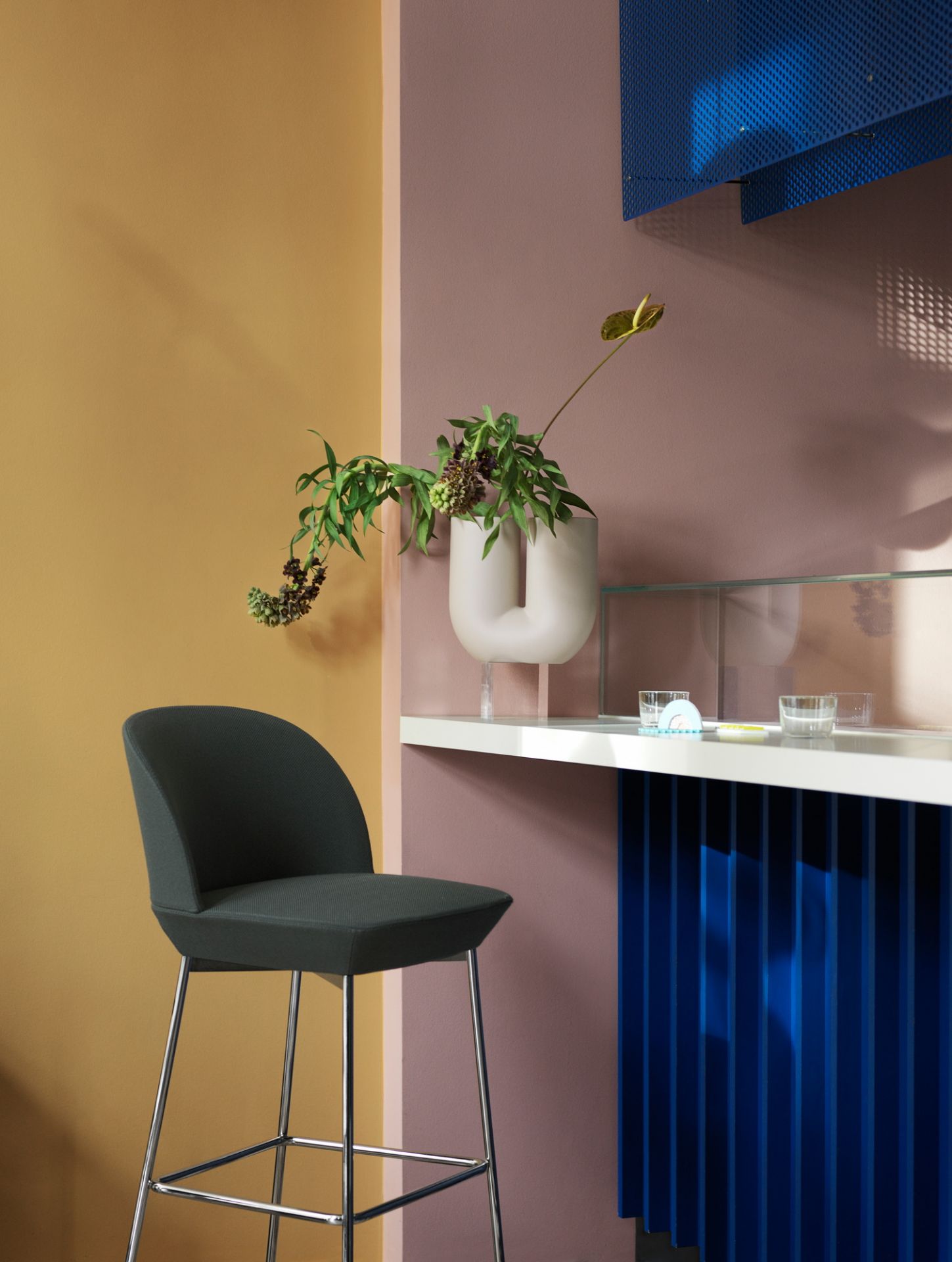 Oslo Bar Stool Barhocker von Muuto, grauer Stoffbezug und verchromtes Gestell vor farbigem Hintergrund.