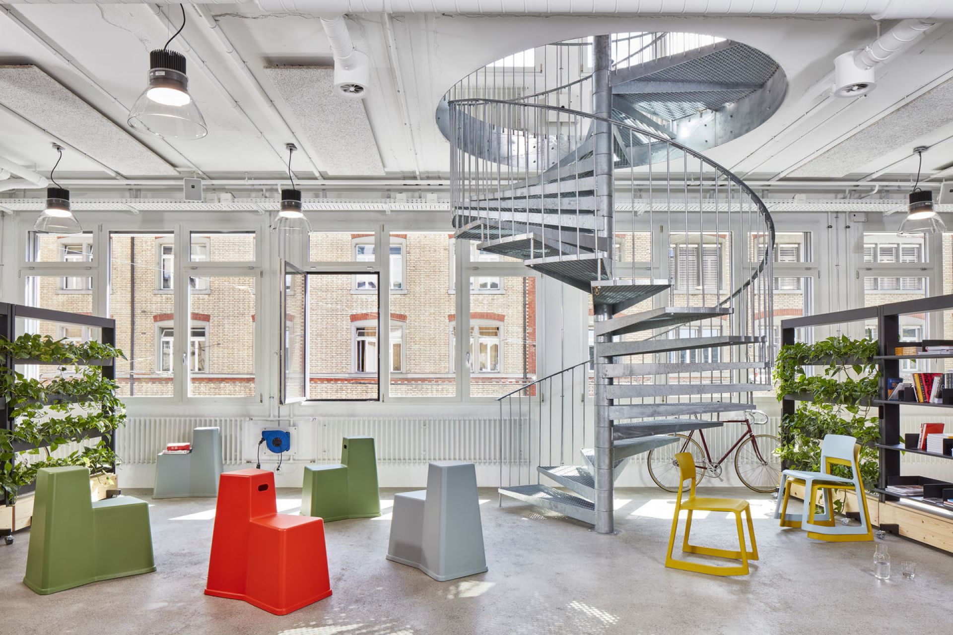 Büro mit bunten Tip Ton Stühlen von Vitra, spiralförmiger Treppe und großen Fenstern für moderne Arbeitsumgebung.