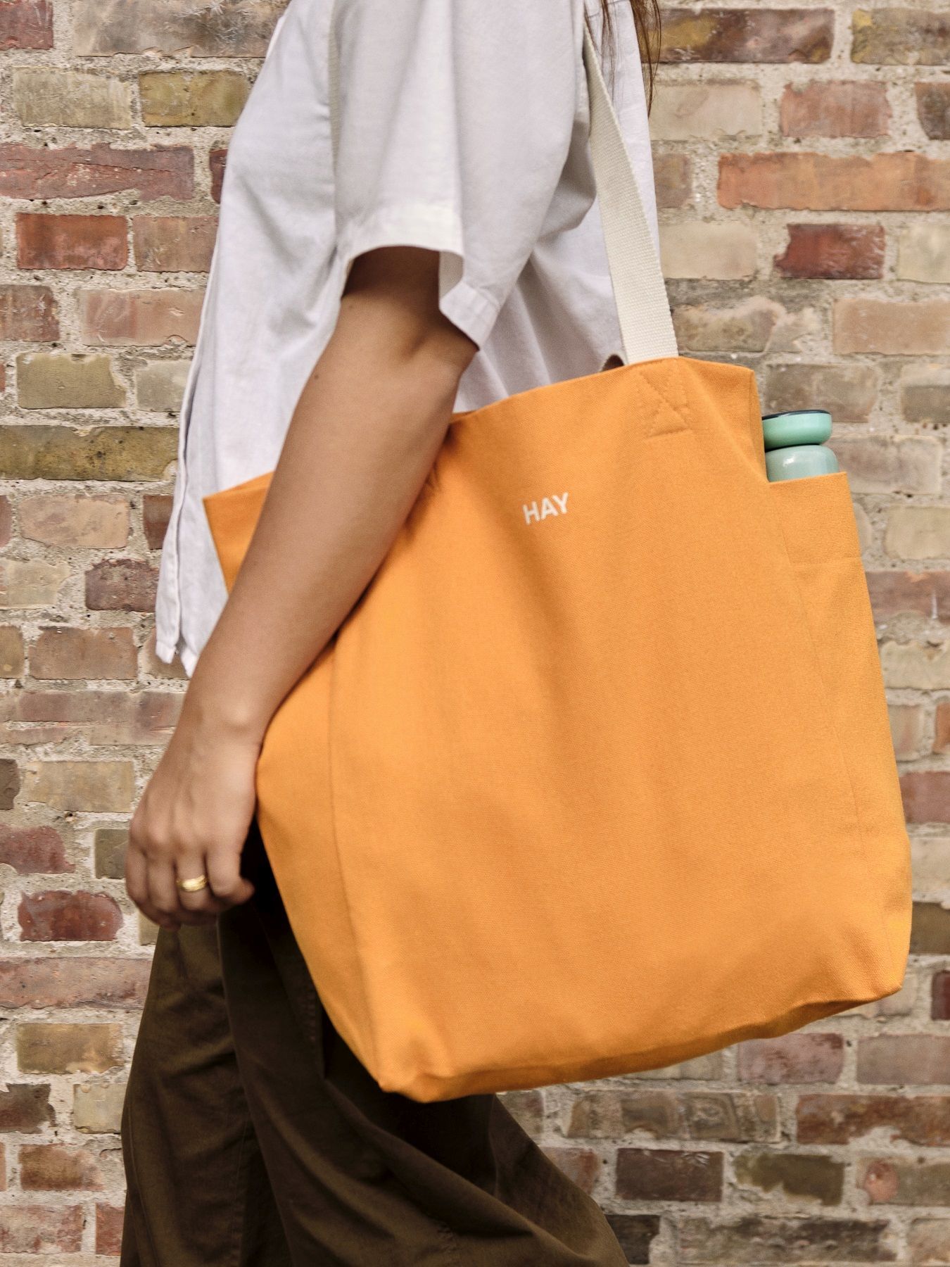 Orangefarbene Hay Everyday Tote Bag aus Stoff, getragen von einer Person vor einer Backsteinmauer.