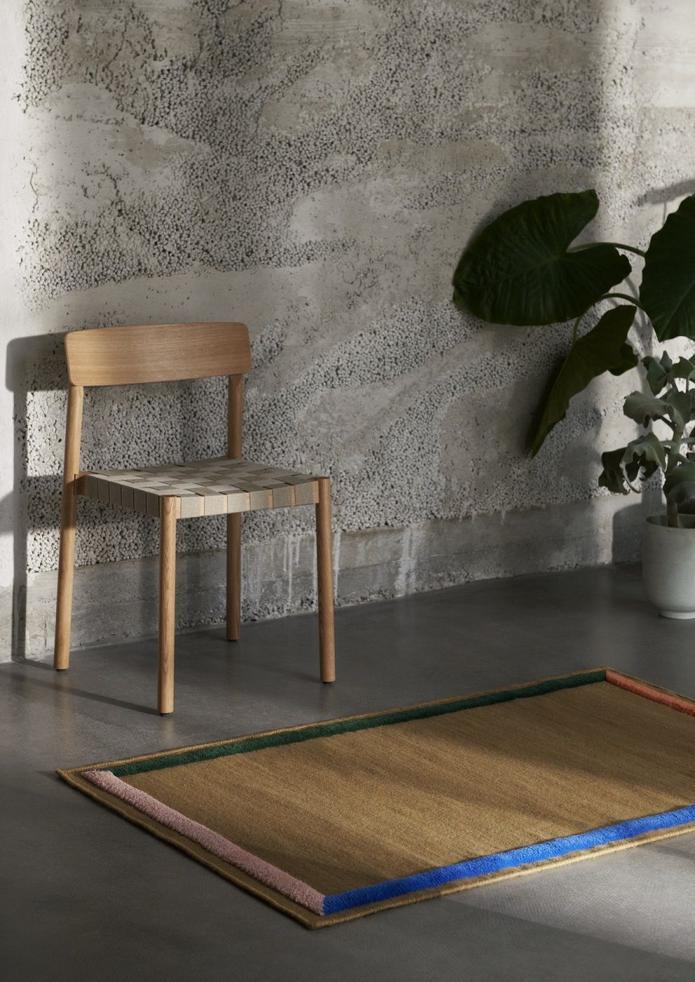 AndTradition Betty TK1 Stuhl aus Holz mit gewebter Sitzfläche vor Betonwand und Teppich.
