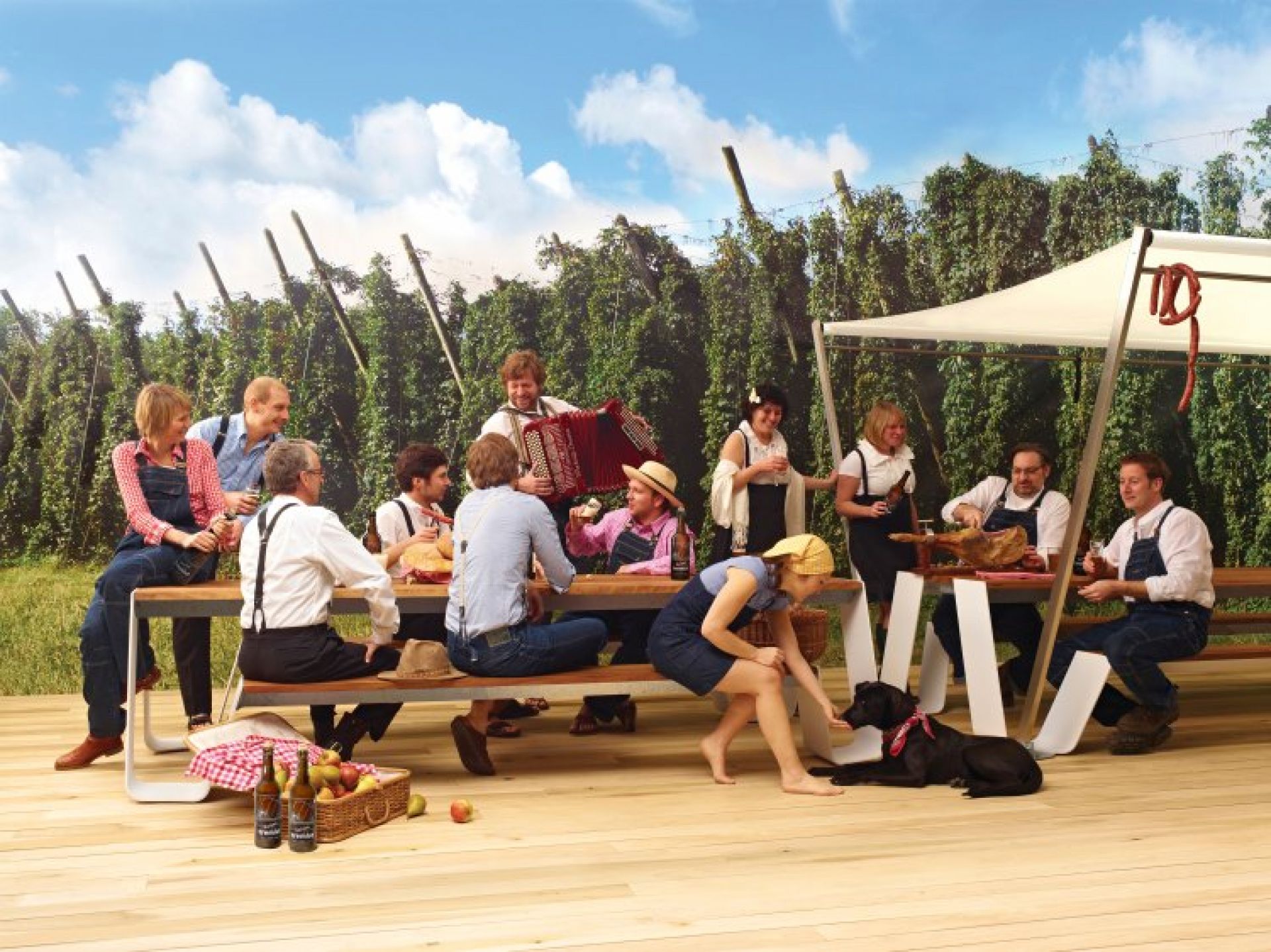 Gruppe genießt Picknick auf Extremis Hopper Picknickbank im Freien mit Speisen, Getränken und Akkordeonmusik vor Hopfenfeld-Hintergrund.