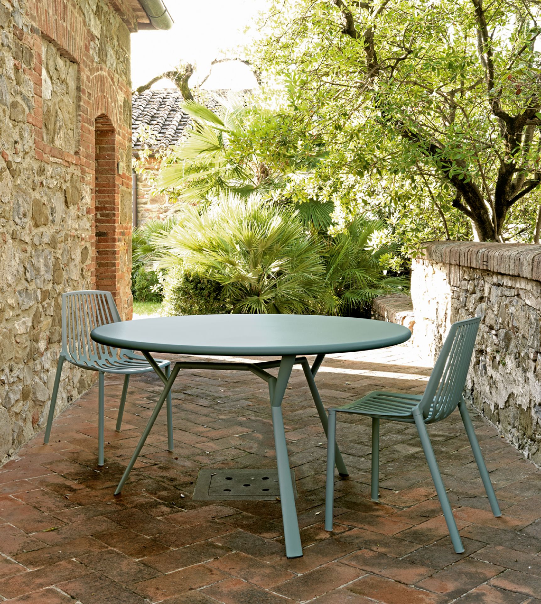 Rion Outdoor Stuhl 2er-Set und runder Tisch auf einer Terrasse mit Backsteinmauer und Pflanzen.
