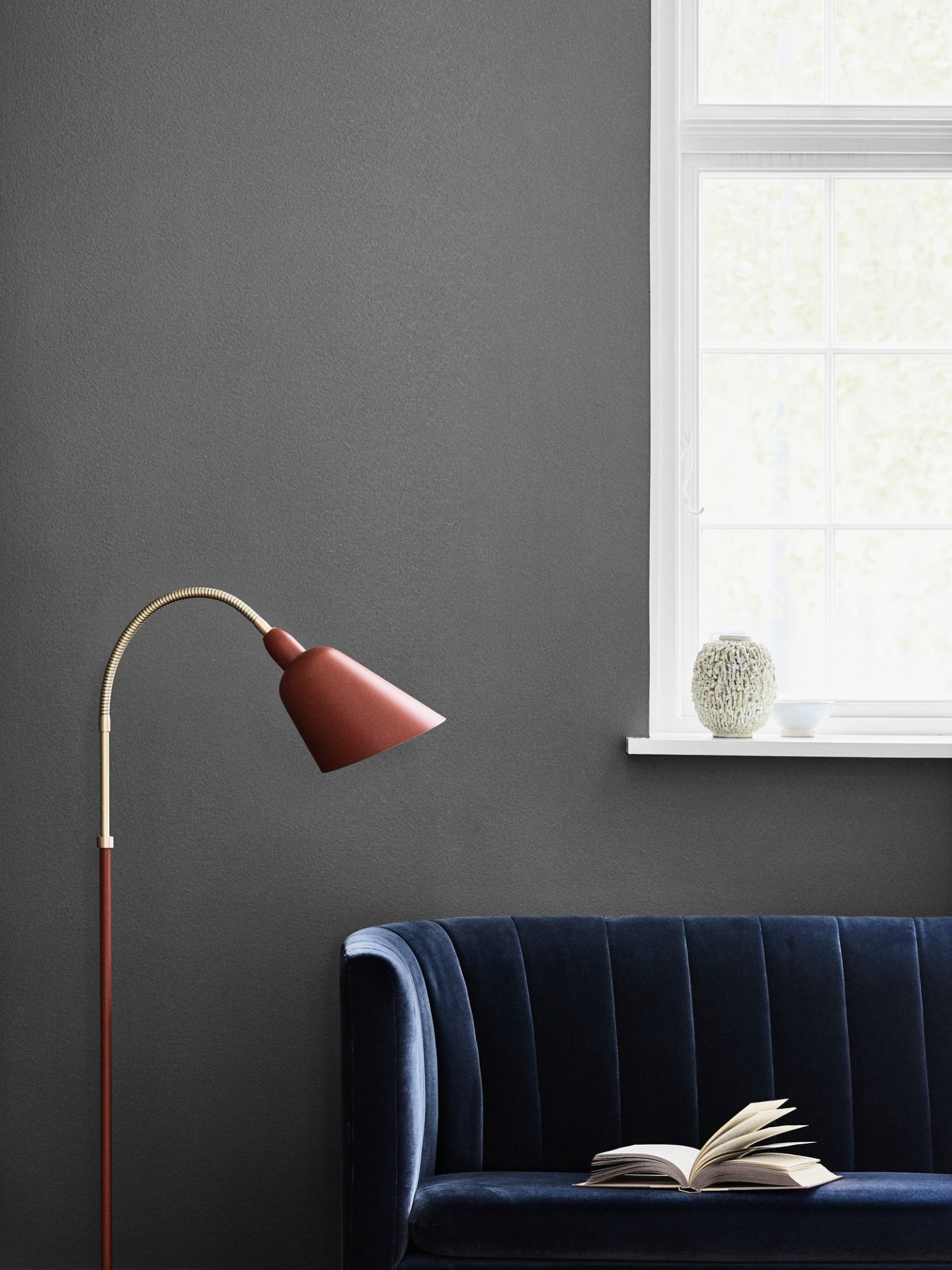 AndTradition Loafer Sofa, blaues Samt Sofa mit Buch und roter Stehlampe vor Fenster.