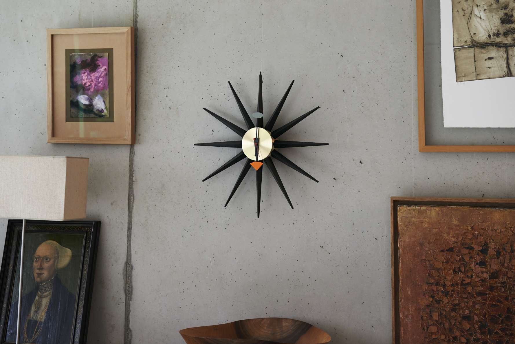 Schwarze Sunburst Clock Wanduhr von Vitra mit Messing-Ziffernblatt an einer grauen Betonwand, umgeben von Kunstwerken.