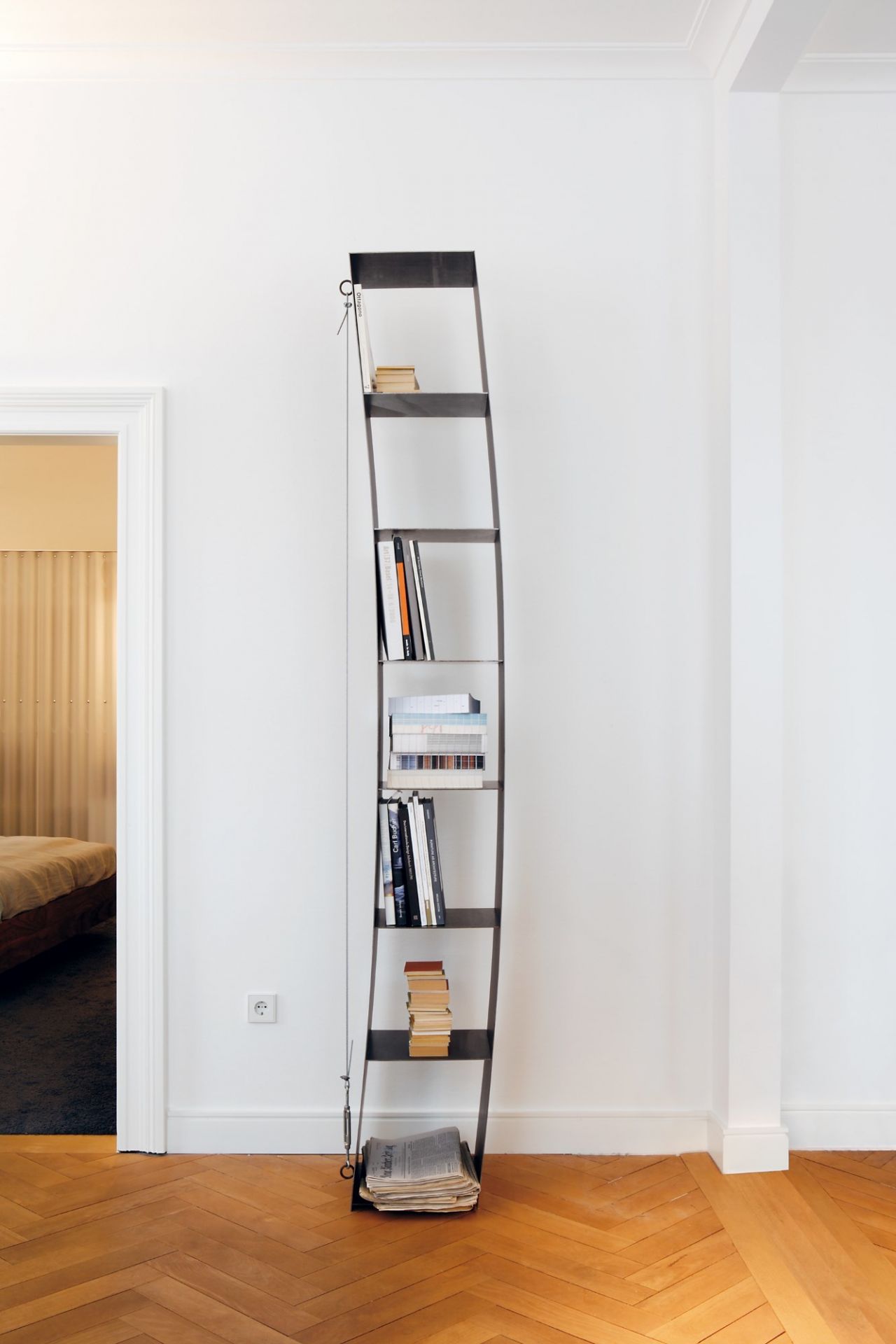 Gespanntes Regal von Moormann, gefüllt mit Büchern und Zeitungen, vor weißer Wand und Parkettboden.