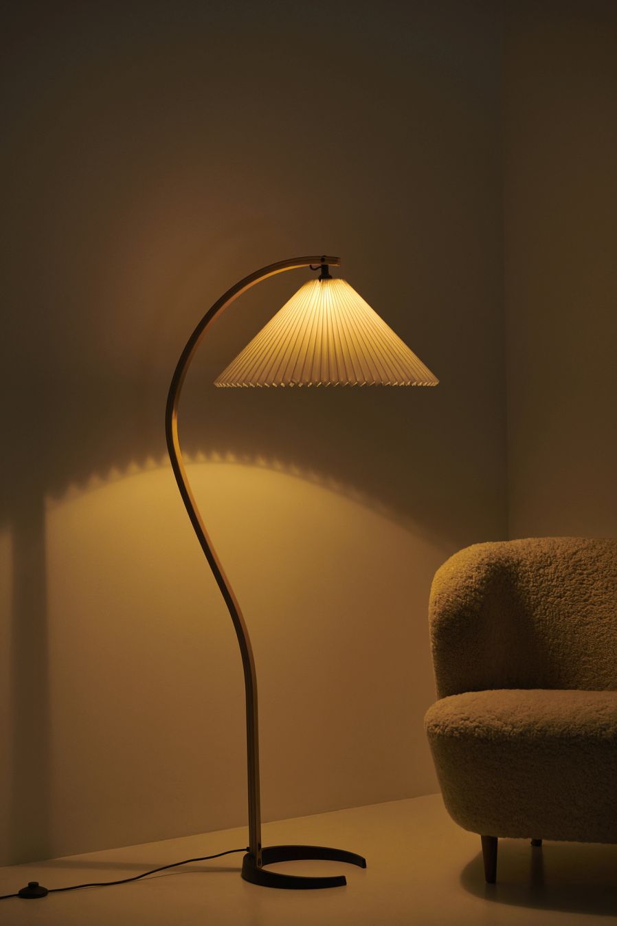 Elegante Timberline Stehleuchte von Gubi mit plissiertem Lampenschirm und warmem Licht neben einem Sessel.