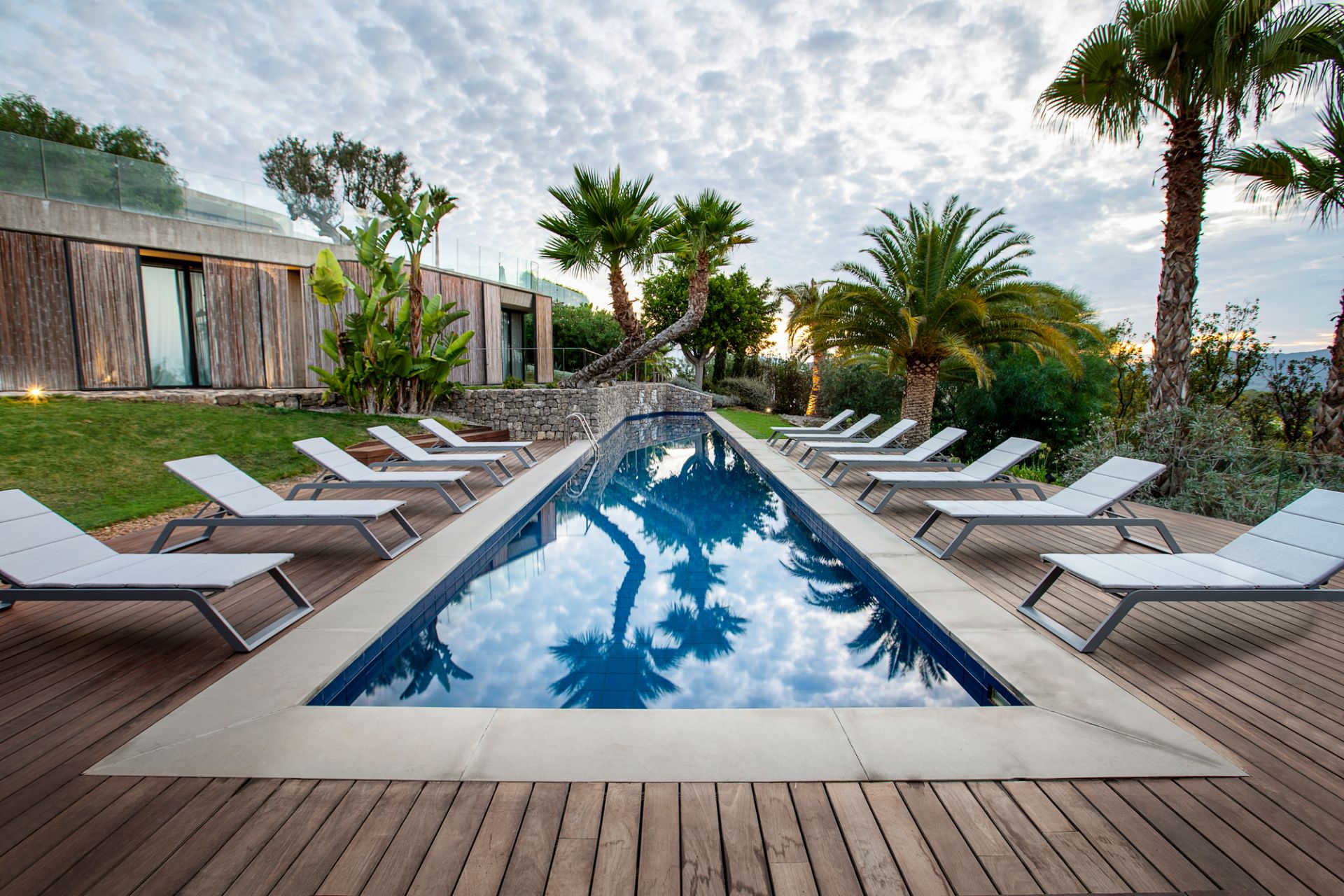 Cane-Line Siesta Sonnenliegen aus grauem Aluminium und Textil am Pool auf Holzdeck mit Palmen.