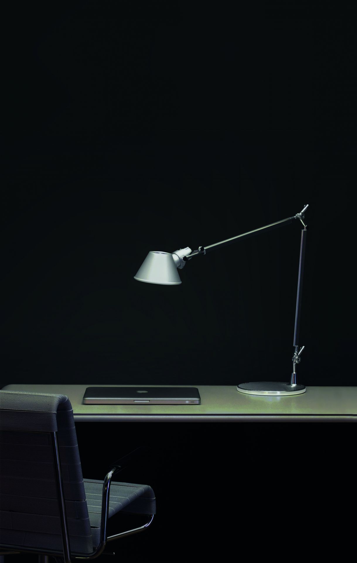 Silberne Artemide Tolomeo Tischleuchte auf Schreibtisch mit Laptop und Bürostuhl vor dunklem Hintergrund.