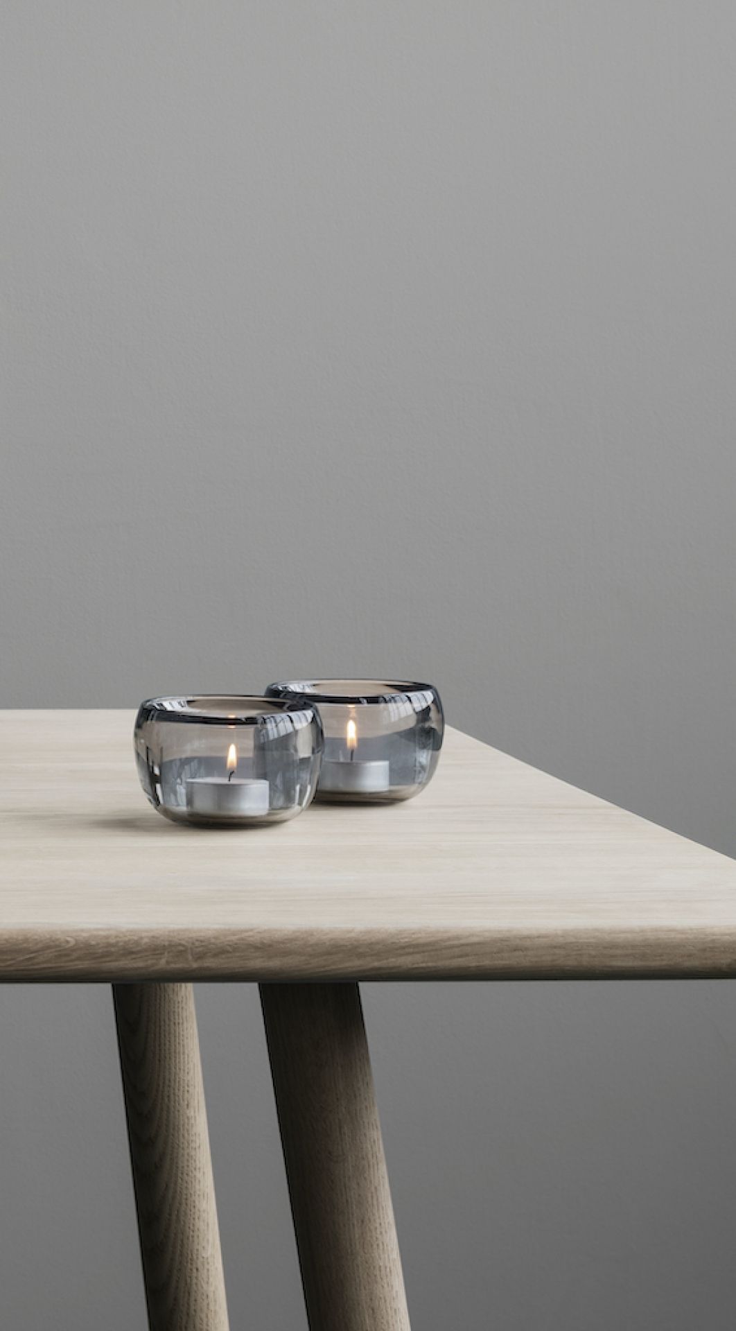 Zwei graue Stelton Ora Teelichthalter aus Glas mit brennenden Teelichtern auf einem Holztisch.