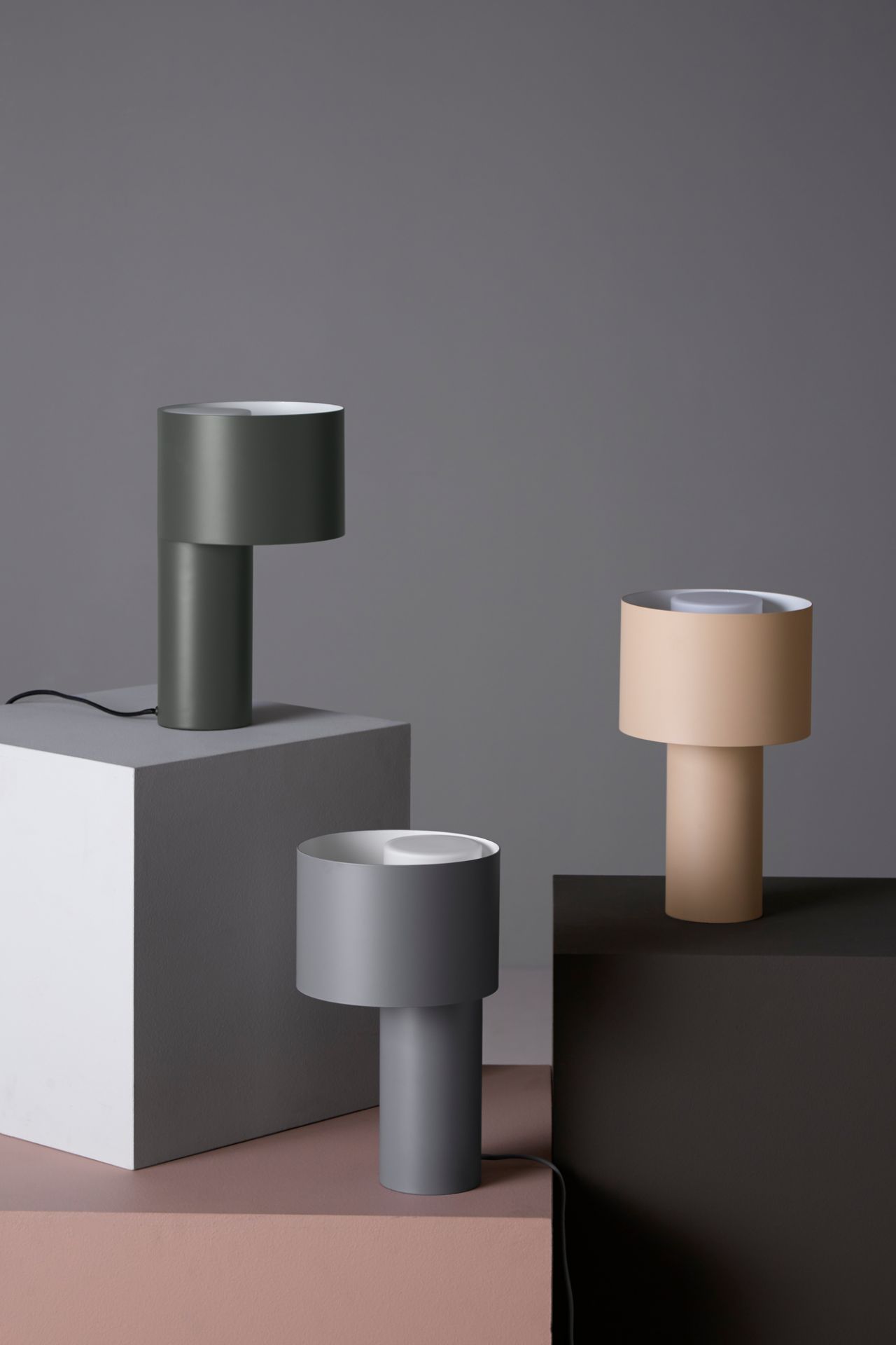 Drei moderne Tangent Tischleuchten von Woud in Grau, Beige und Olivgrün auf geometrischen Blöcken.