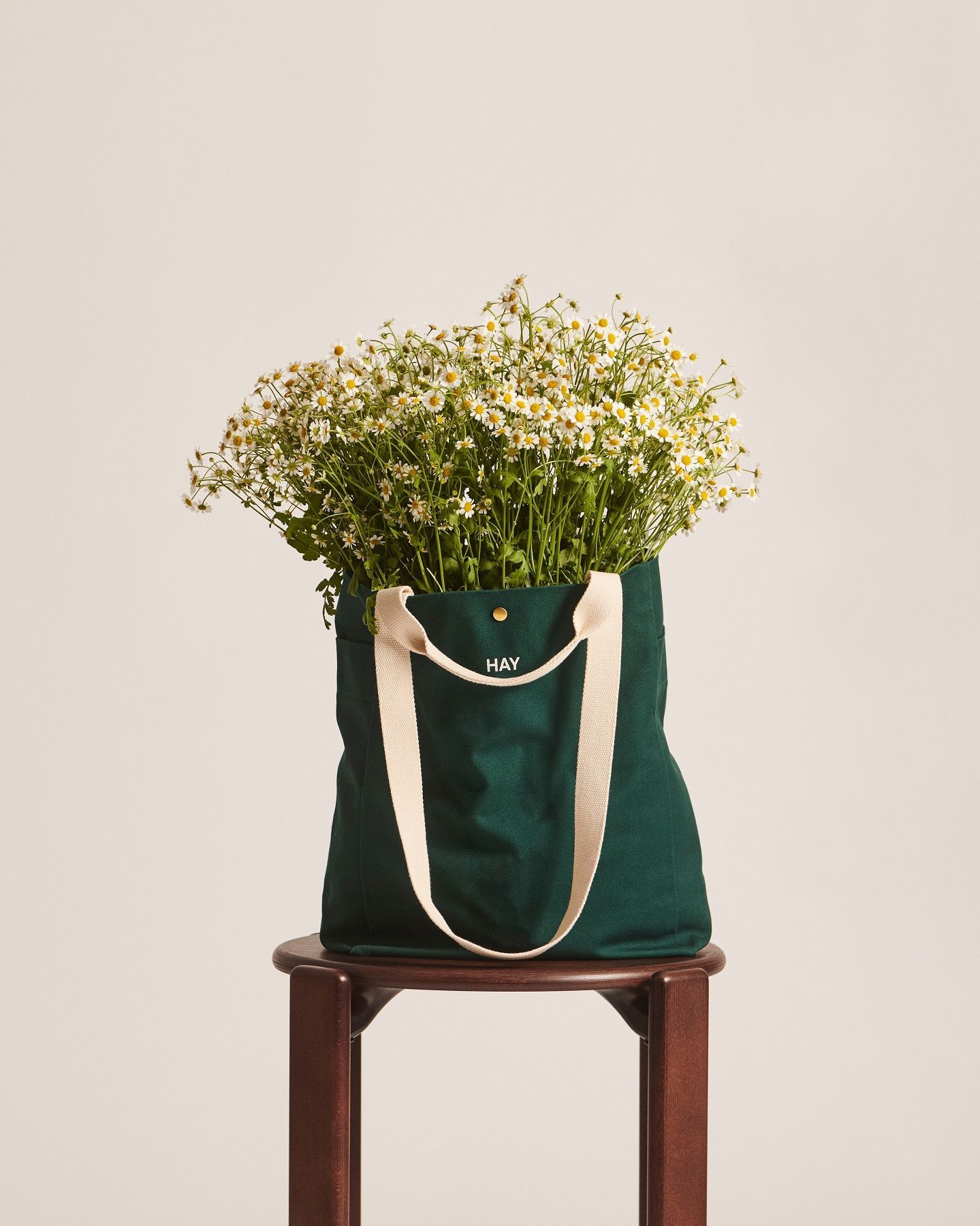 Grüne Hay Everyday Tote Bag auf einem Hocker, gefüllt mit einem üppigen Strauß Gänseblümchen.