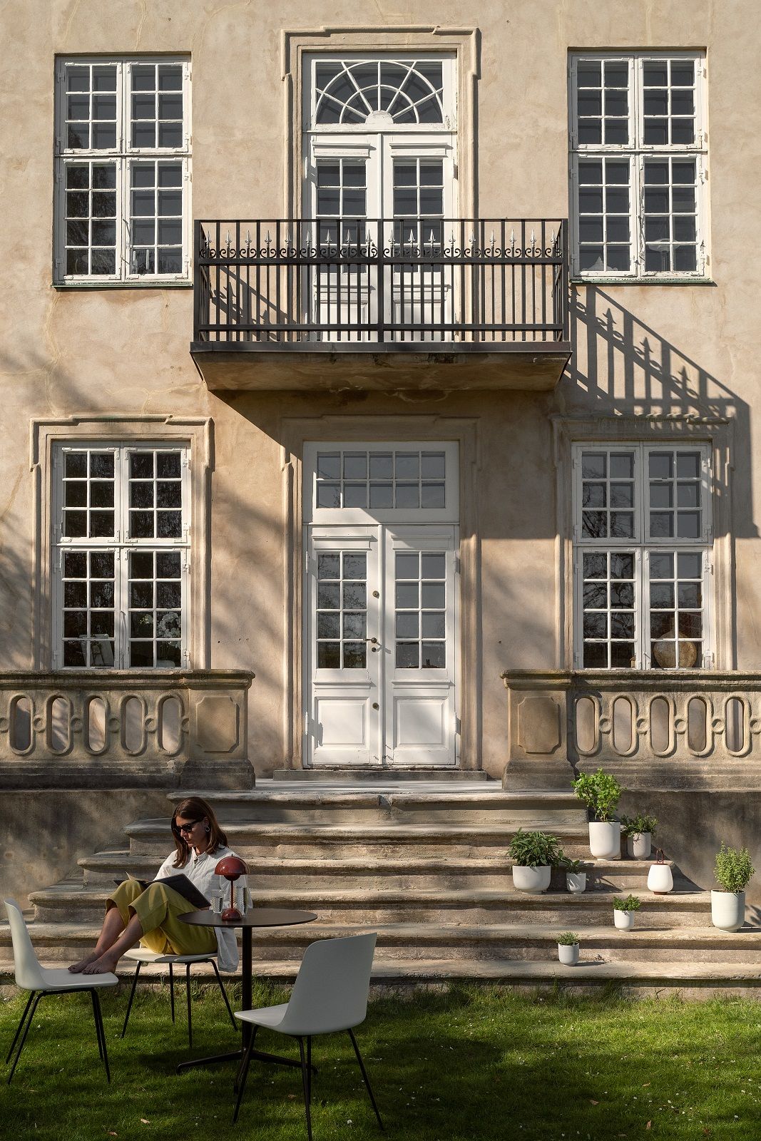 Frau liest auf den Stufen eines Hauses, sitzend auf AndTradition Rely HW70 Outdoor Stühlen mit Gartentisch.