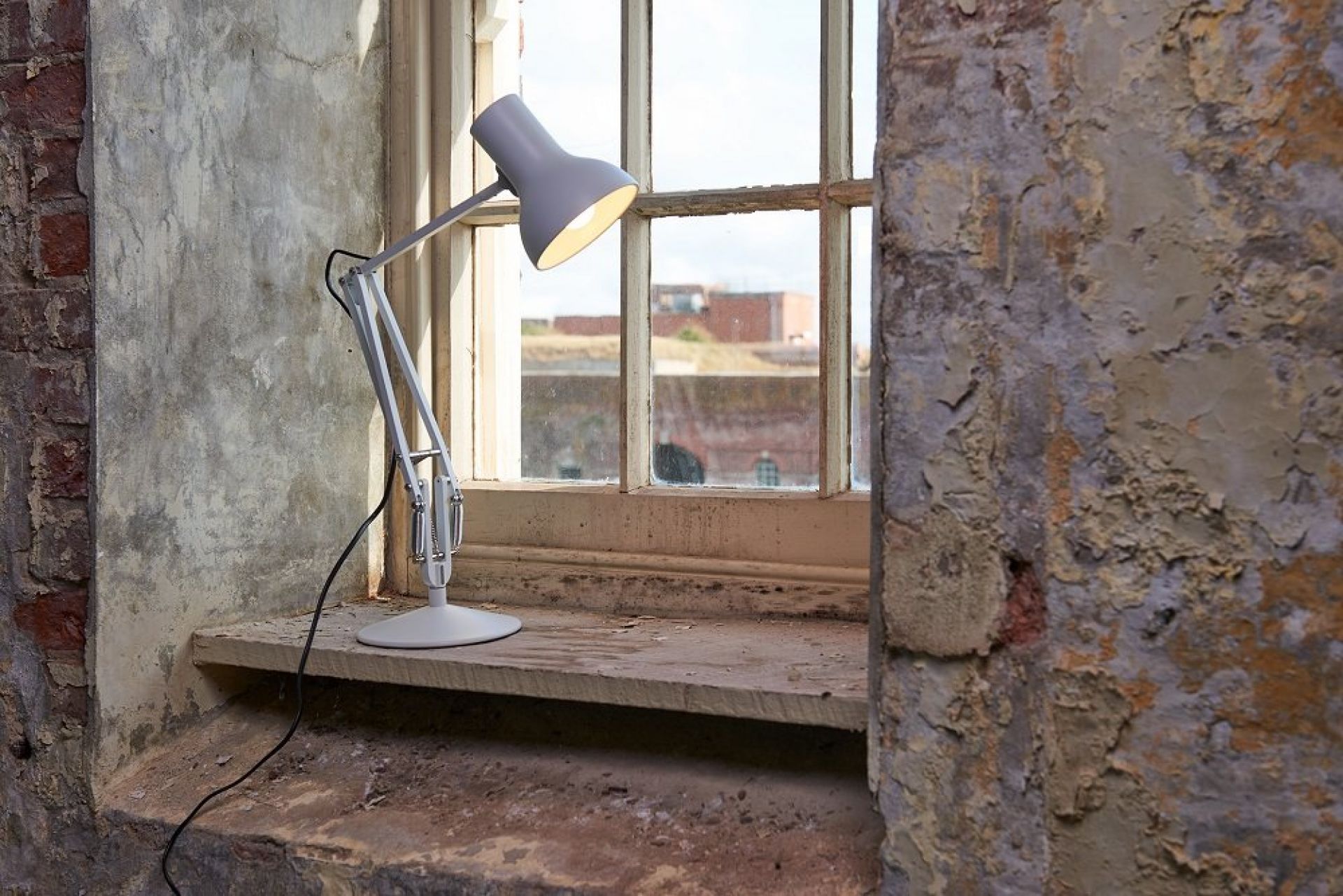 Graue Anglepoise Type 75 Mini Tischleuchte auf einer Fensterbank vor einem alten Fenster.