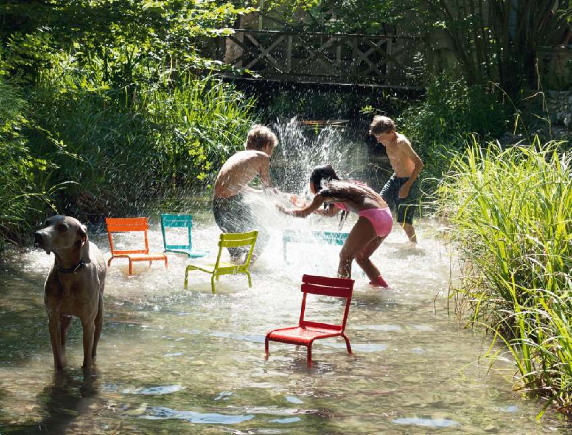 Kinder spielen im Wasser mit bunten Luxembourg Kinderstühlen von Fermob, ein Hund steht im Vordergrund.