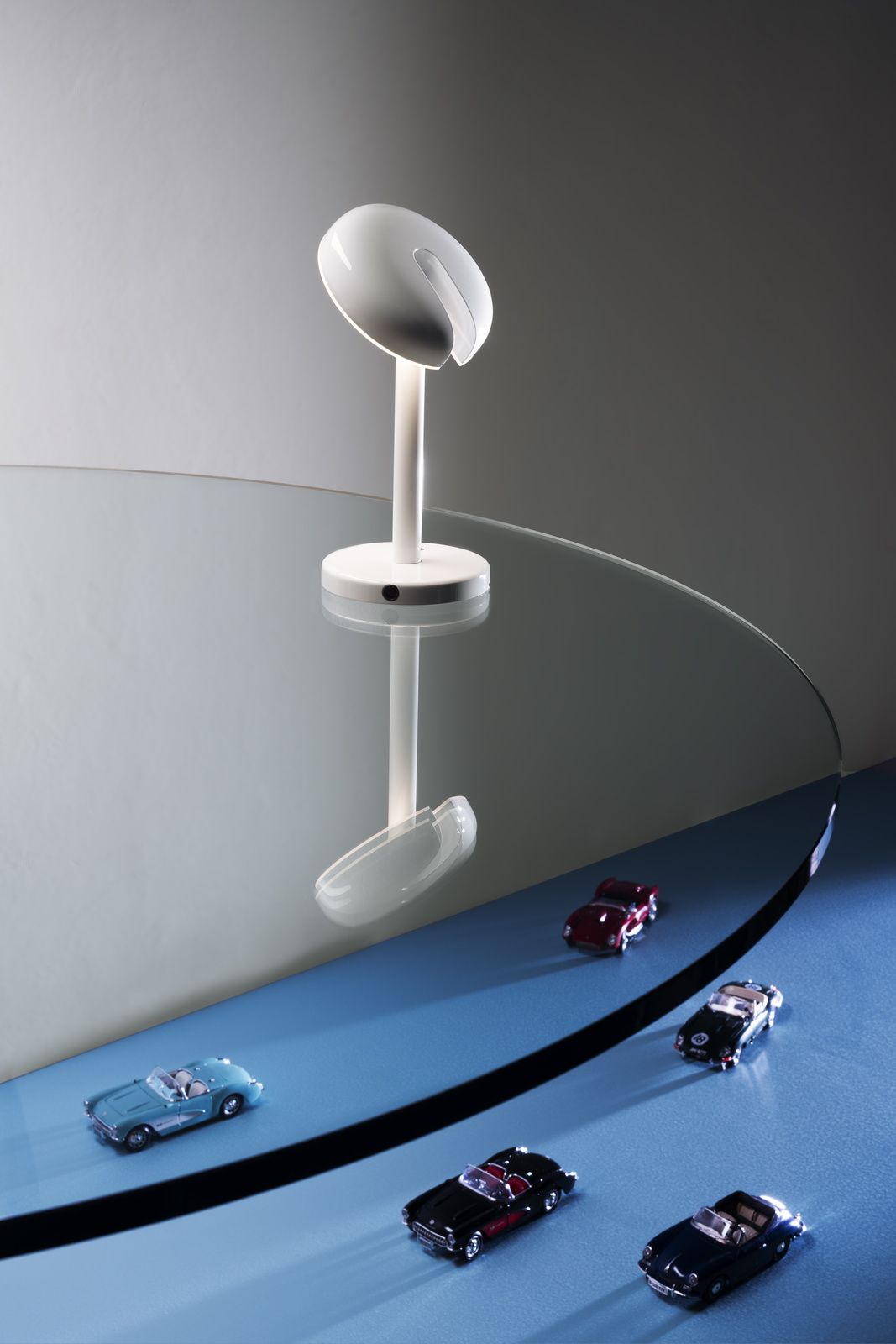Weiße Martinelli Luce Tischleuchte "Cabriolette" auf Glastisch mit Spiegelung und Spielzeugautos im Vordergrund.