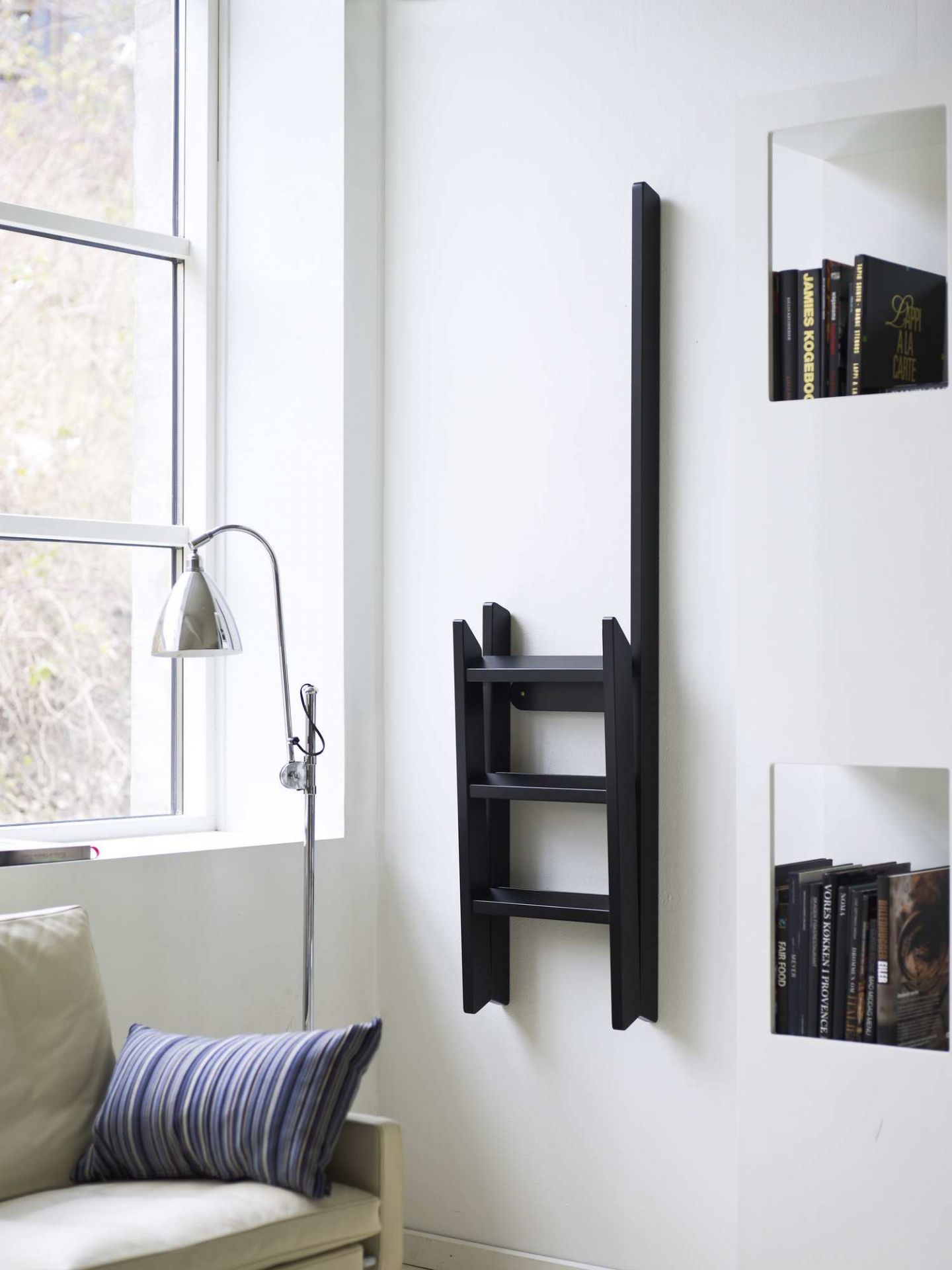 Schwarze Hindevadgaard Trittleiter an weißer Wand, zusammengeklappt als Dekoelement, modernes Wohnzimmer mit Fenster und Lampe.