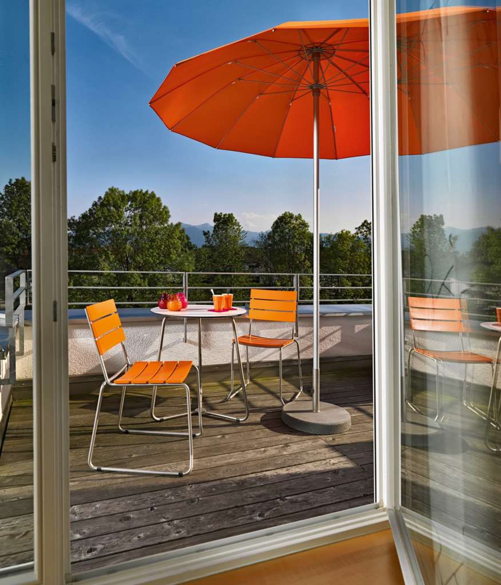 Weishäupl Balcony Bistrotisch mit Edelstahlgestell und orangefarbenen Stühlen auf einem sonnigen Balkon mit Sonnenschirm.