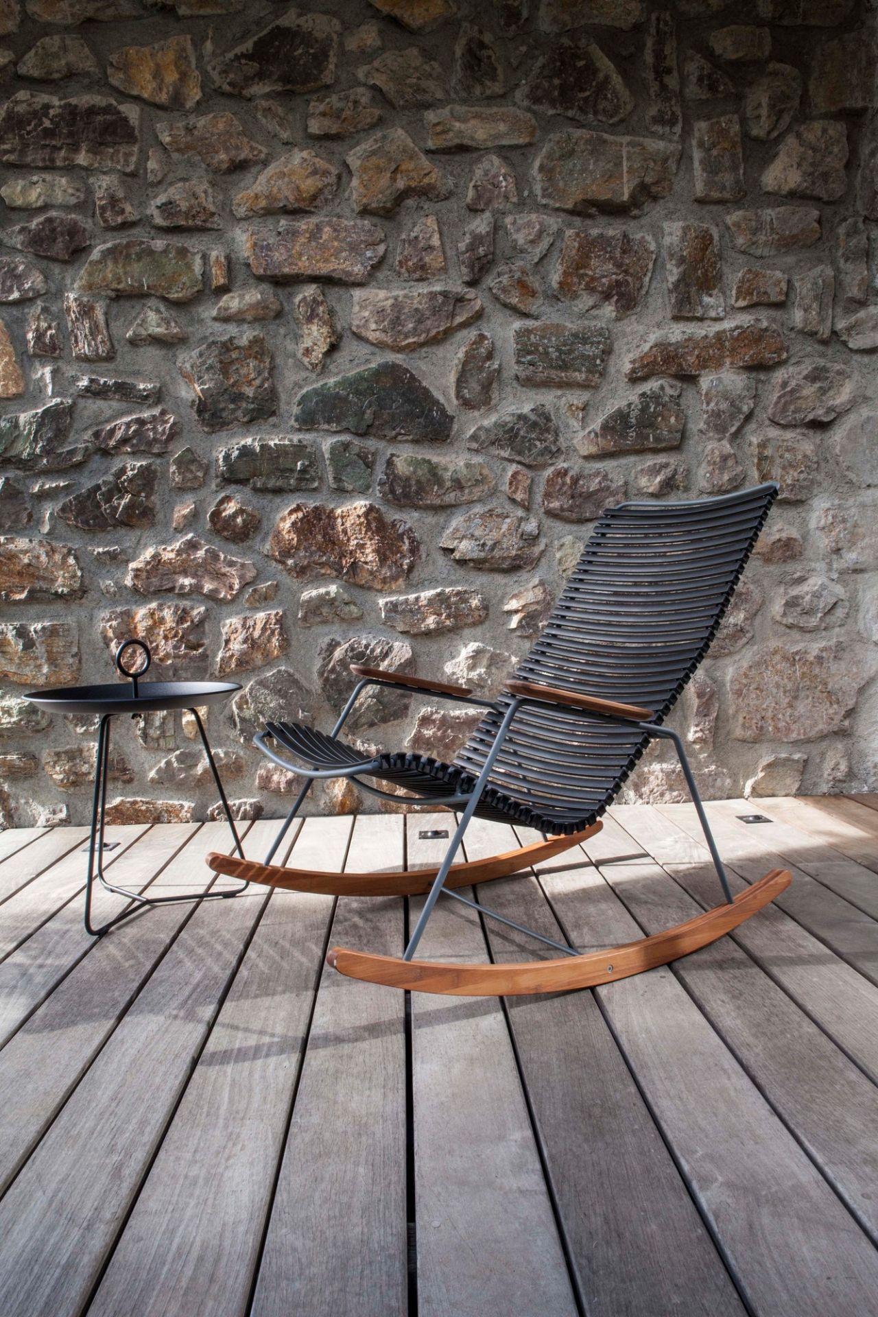 Houe Click Schaukelstuhl für den Außenbereich mit Beistelltisch auf Holzterrasse vor Steinwand.