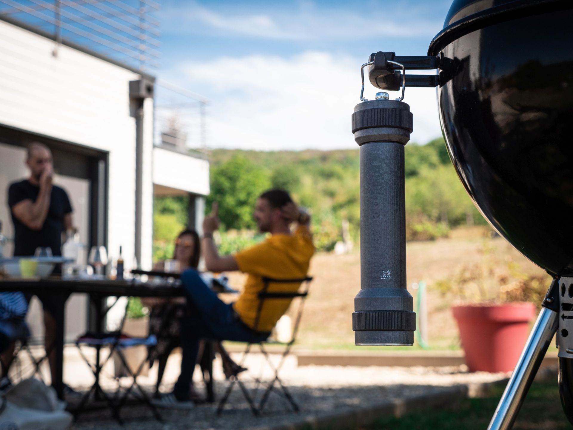 Graphitfarbene Peugeot BBQ Pfeffermühle am Grill befestigt, im Hintergrund eine entspannte Grillrunde im Garten.