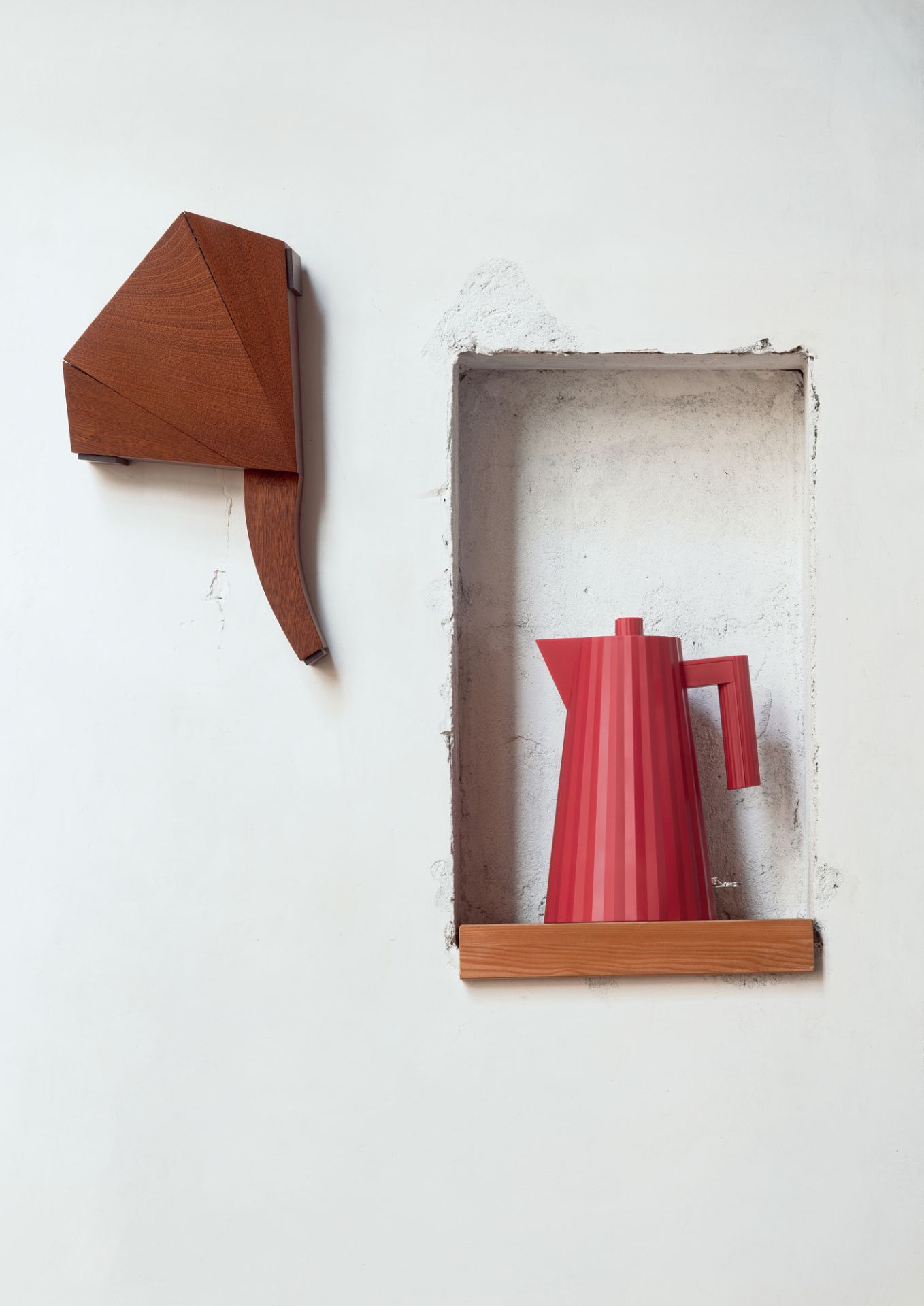 Roter Alessi Plissé Wasserkocher in Nische, daneben Holzobjekt an weißer Wand. Design Wasserkocher.