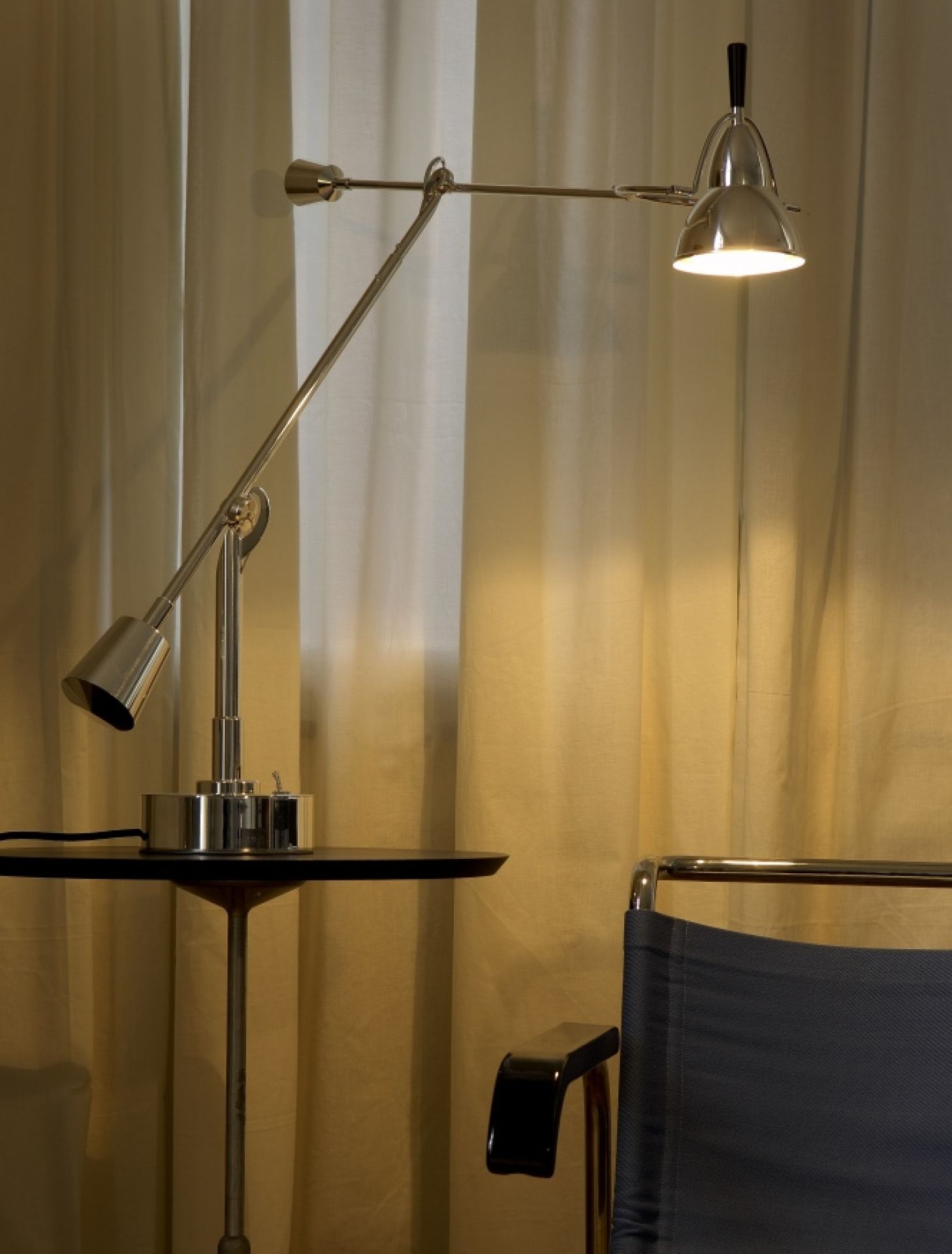 Moderne, verstellbare Schreibtischlampe aus Chrom mit schwarzem Griff auf einem Beistelltisch, vor Vorhängen.