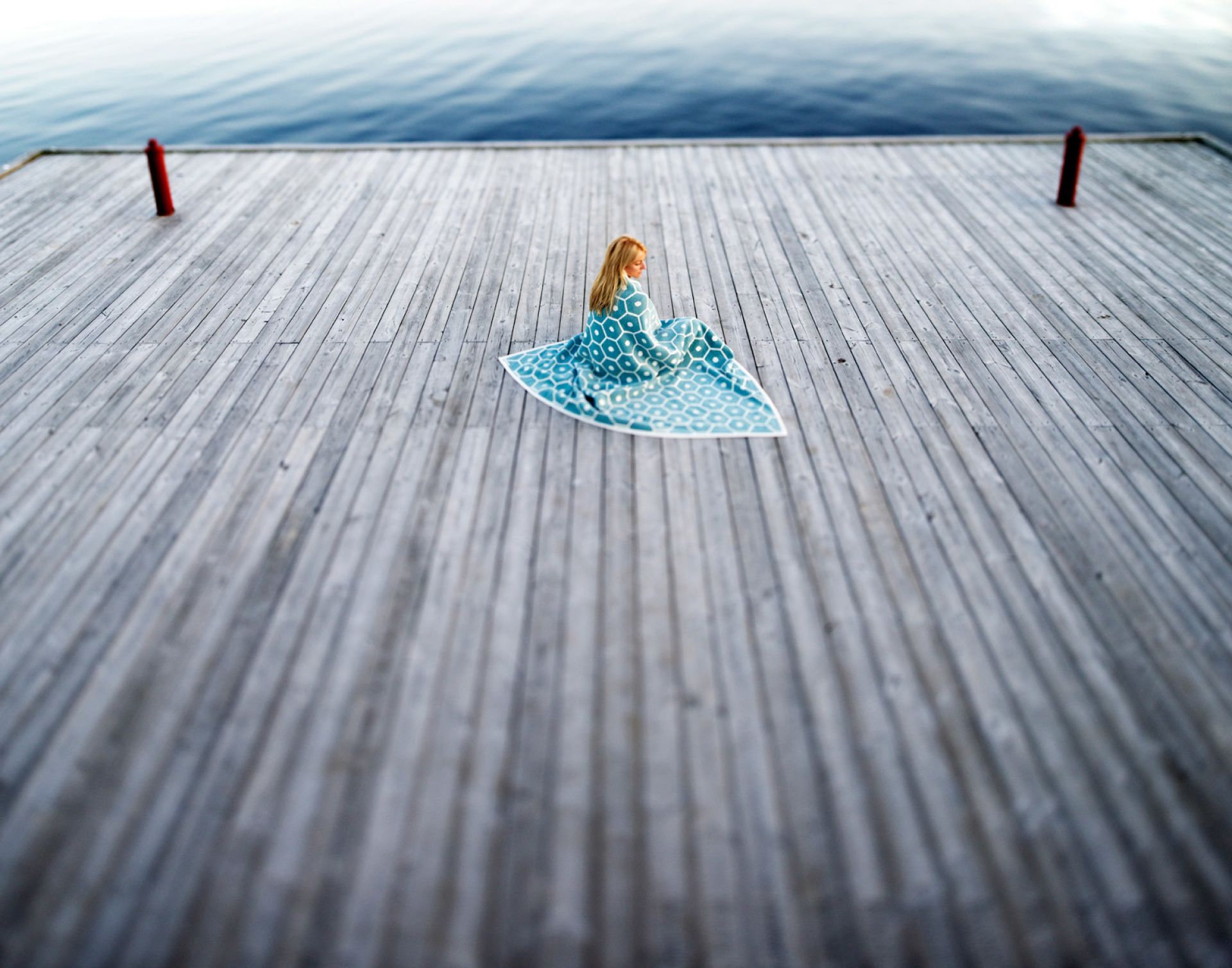 Frau sitzt auf Holzsteg, gehüllt in eine blaue Pappelina Wolldecke mit Muster.
