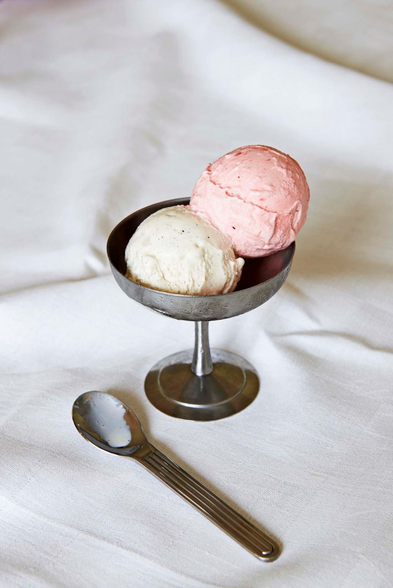 Zwei Eiskugeln in einem silbernen Eisbecher (Italian Ice Cup) mit Löffel auf weißem Stoff.