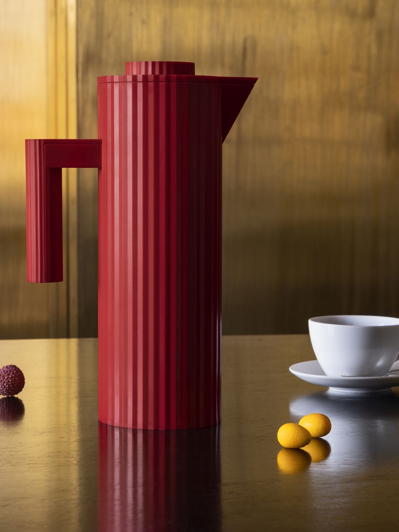 Rote Alessi Isolierkanne mit Plissee-Design neben Tasse und Früchten auf glänzender Oberfläche.