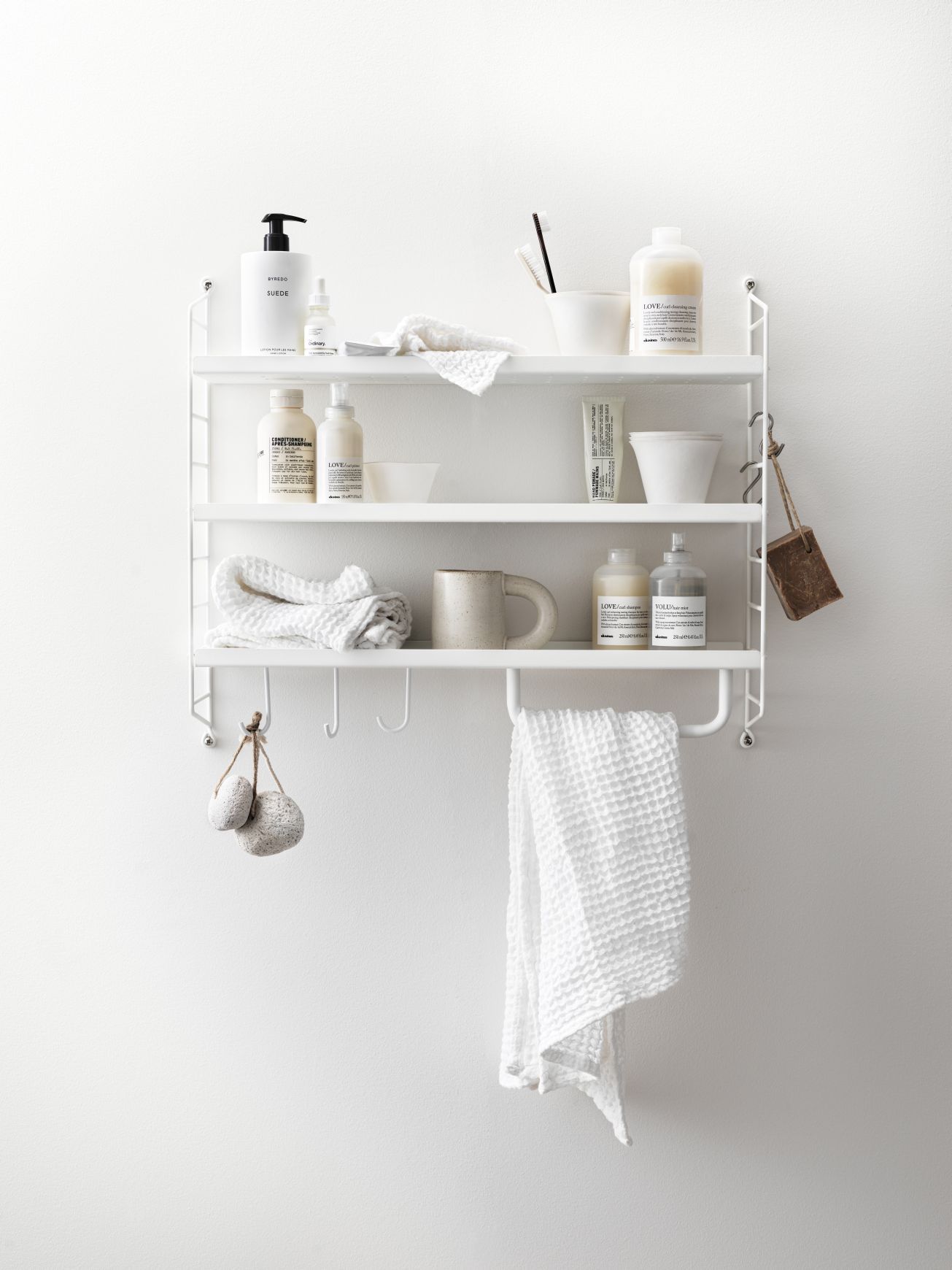 Weißes String Pocket Regal aus Metall mit Badartikeln und Handtüchern an einer weißen Wand.