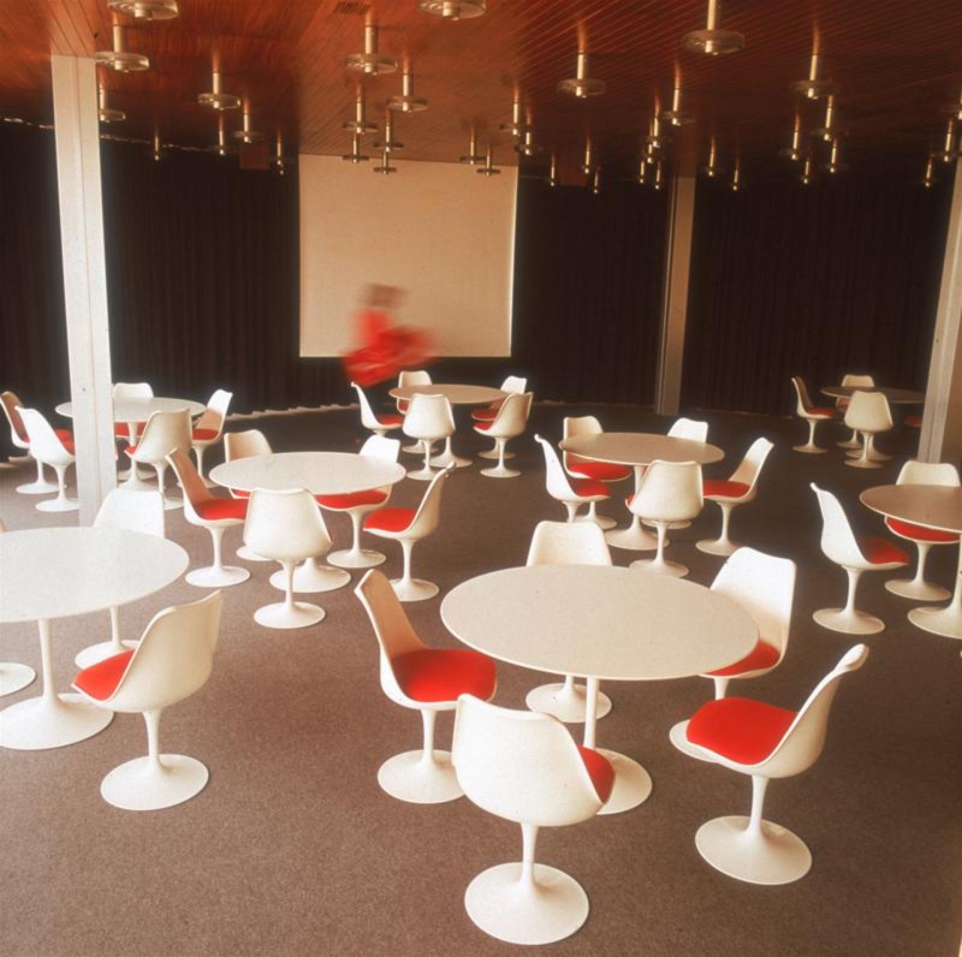 Innenaufnahme mit vielen runden Saarinen Esstischen von Knoll International mit weißem Untergestell und roten Stühlen.