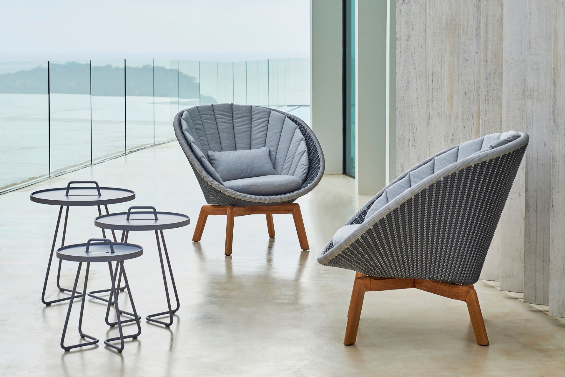 Zwei graue Cane-Line Peacock Loungesessel aus Rattan mit Kissen und drei graue Beistelltische im Außenbereich.