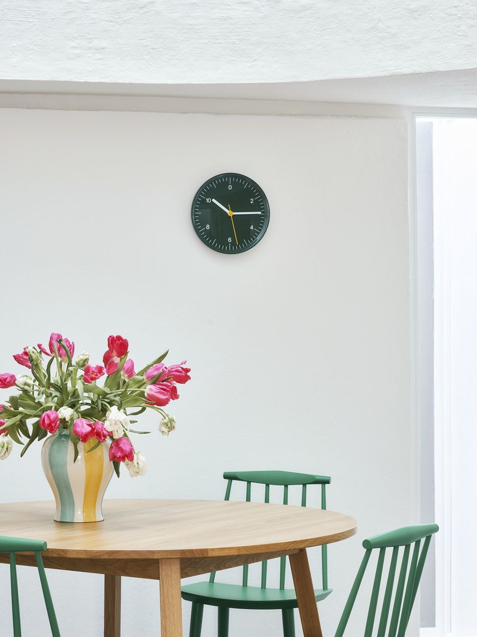 Nahaufnahme der Hay Wanduhr in Schwarz über einem Esstisch mit Blumen und grünen Stühlen.