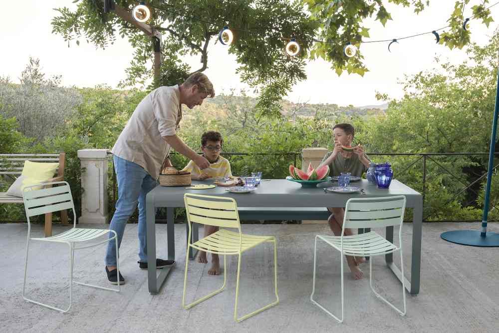 Somerset Bank Outdoor Fermob: Familie genießt Mahlzeit am grauen Gartentisch mit bunten Stühlen.