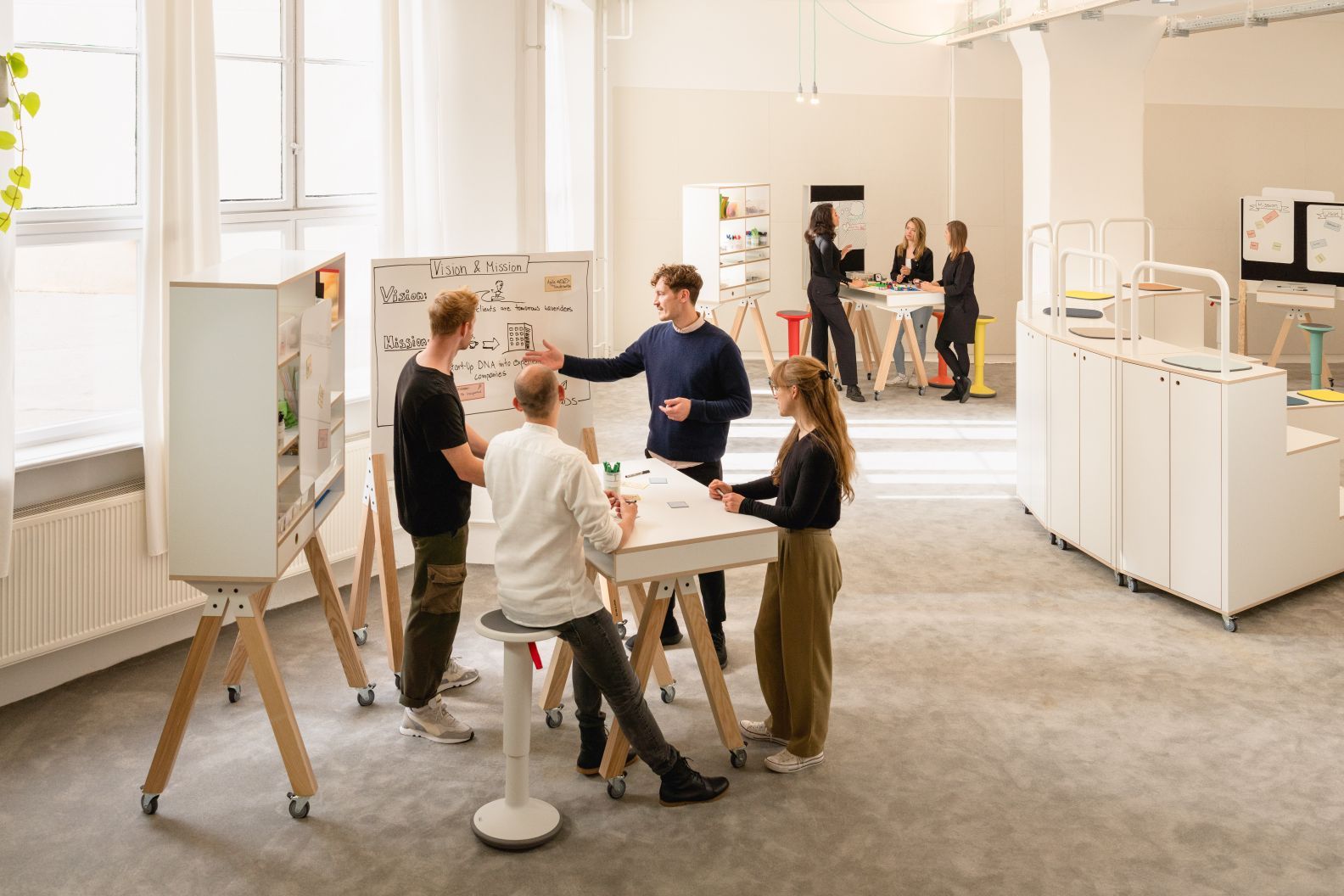 What if We Fly Taskboard: Menschen arbeiten an Whiteboard Medienwand und nutzen Interstuhl Hocker im modernen Büro.