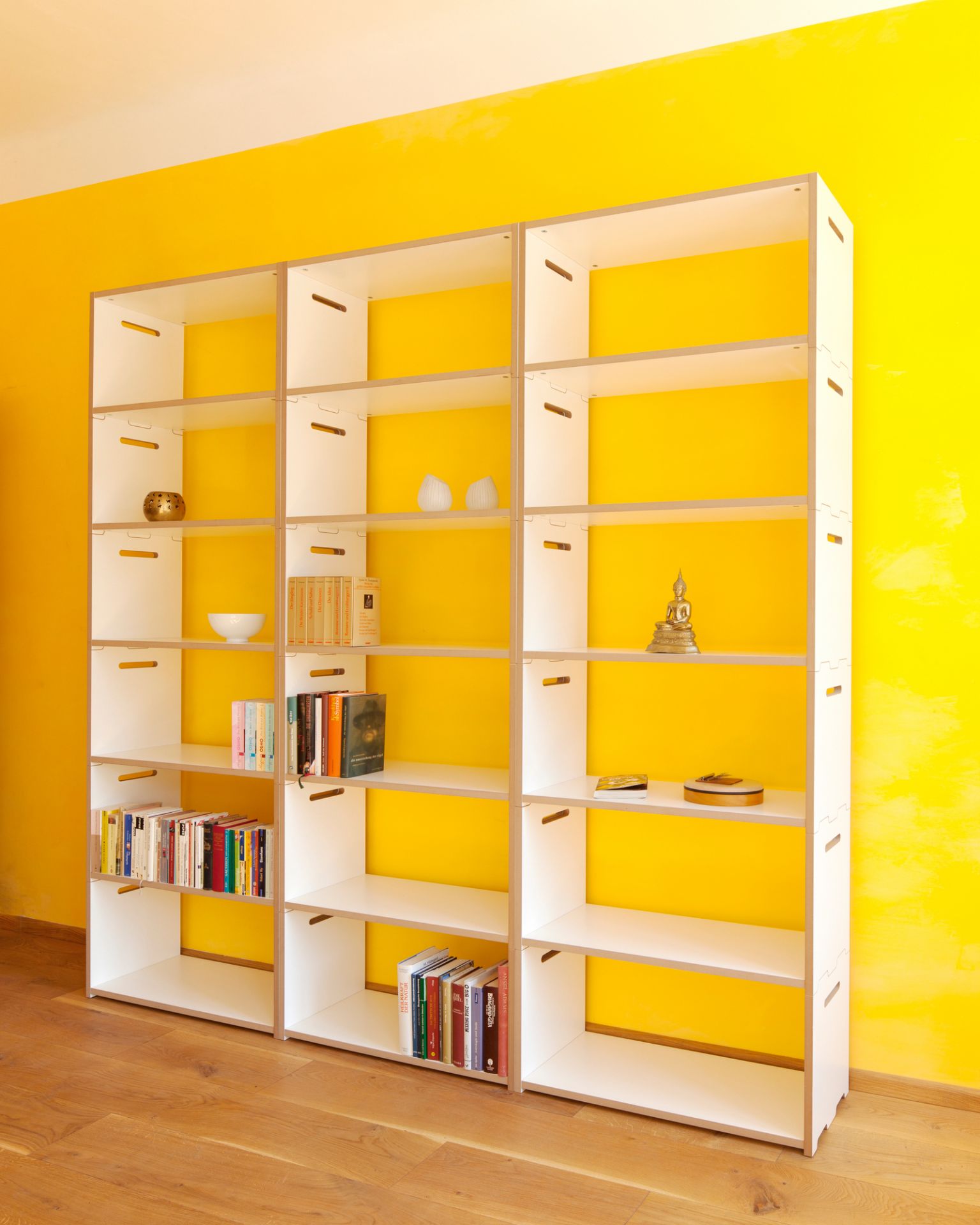 Tojo Hochstapler Regal Grundmodul, weißes modulares Regalsystem vor gelber Wand, Bücher und Dekoration.