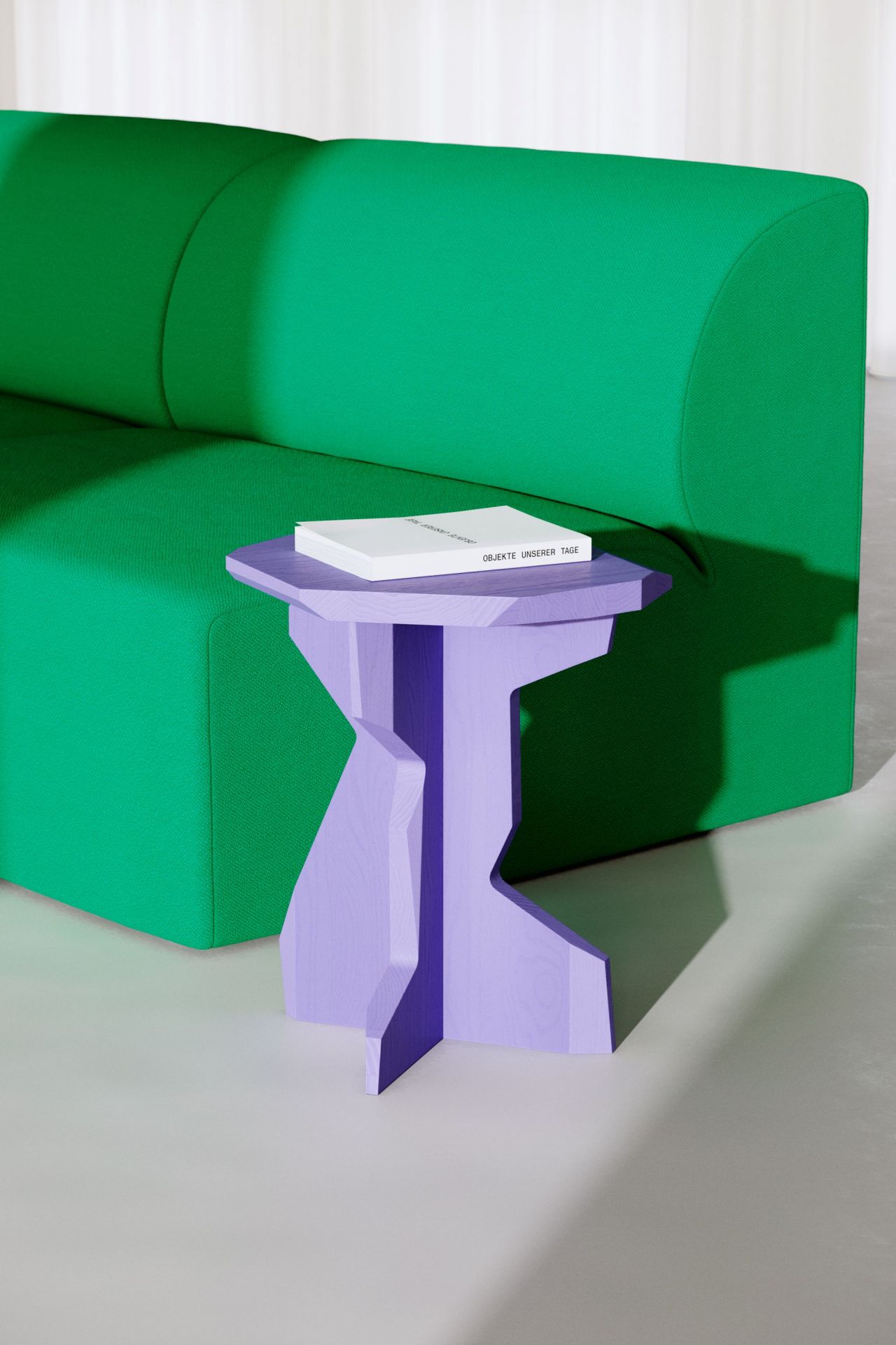 FELS Hocker Esche Out in Lila neben einem grünen Sofa mit einem Buch darauf.