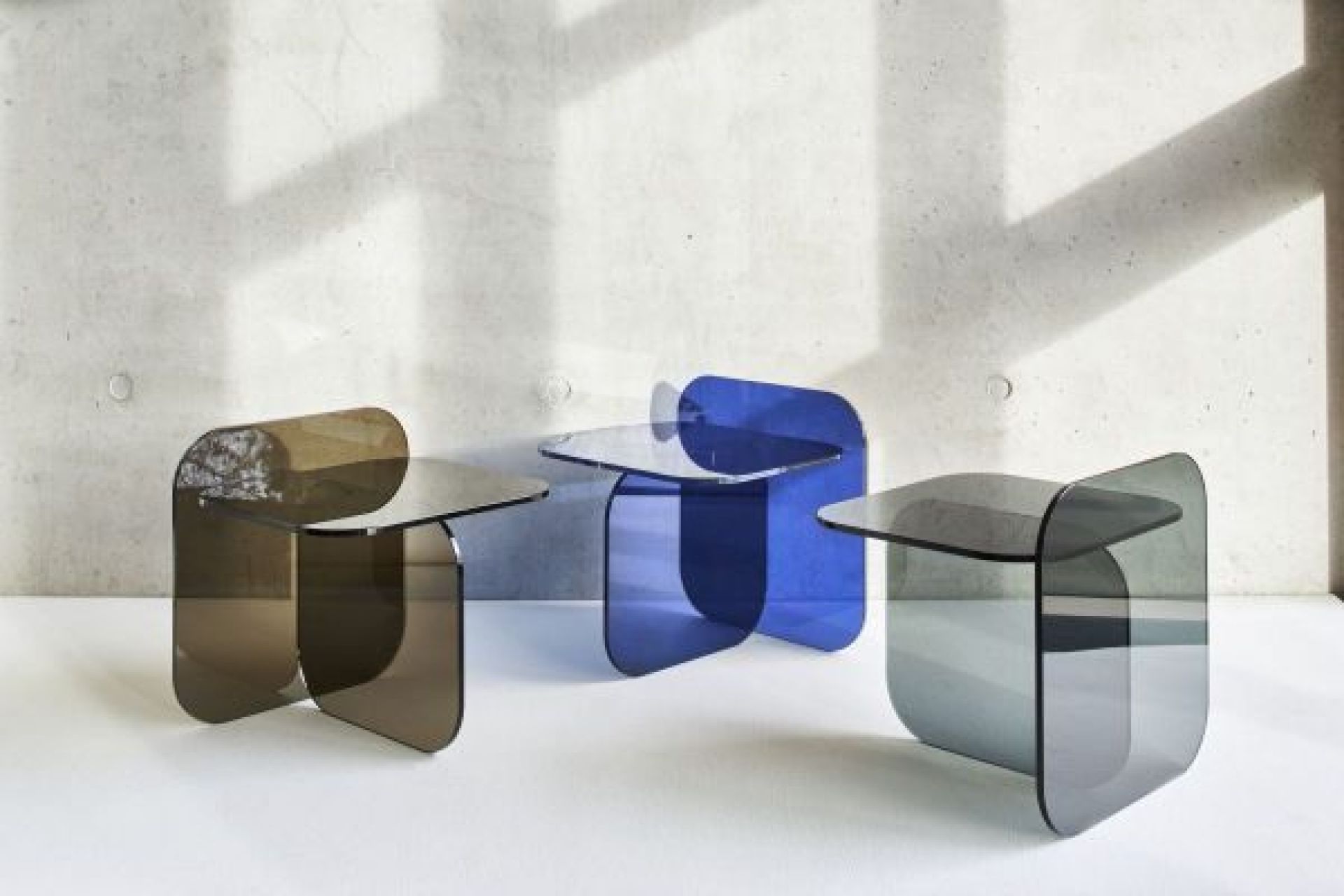 Drei ClassiCon Sol Beistelltische aus Kristallglas in Braun, Grau und Royalblau vor Betonwand.