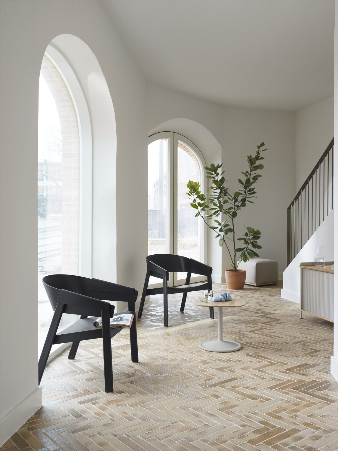 Elegantes Interieur mit zwei schwarzen Muuto Sesseln, kleinem Tisch und Pflanze, im Hintergrund ein Echo Pouf Hocker.