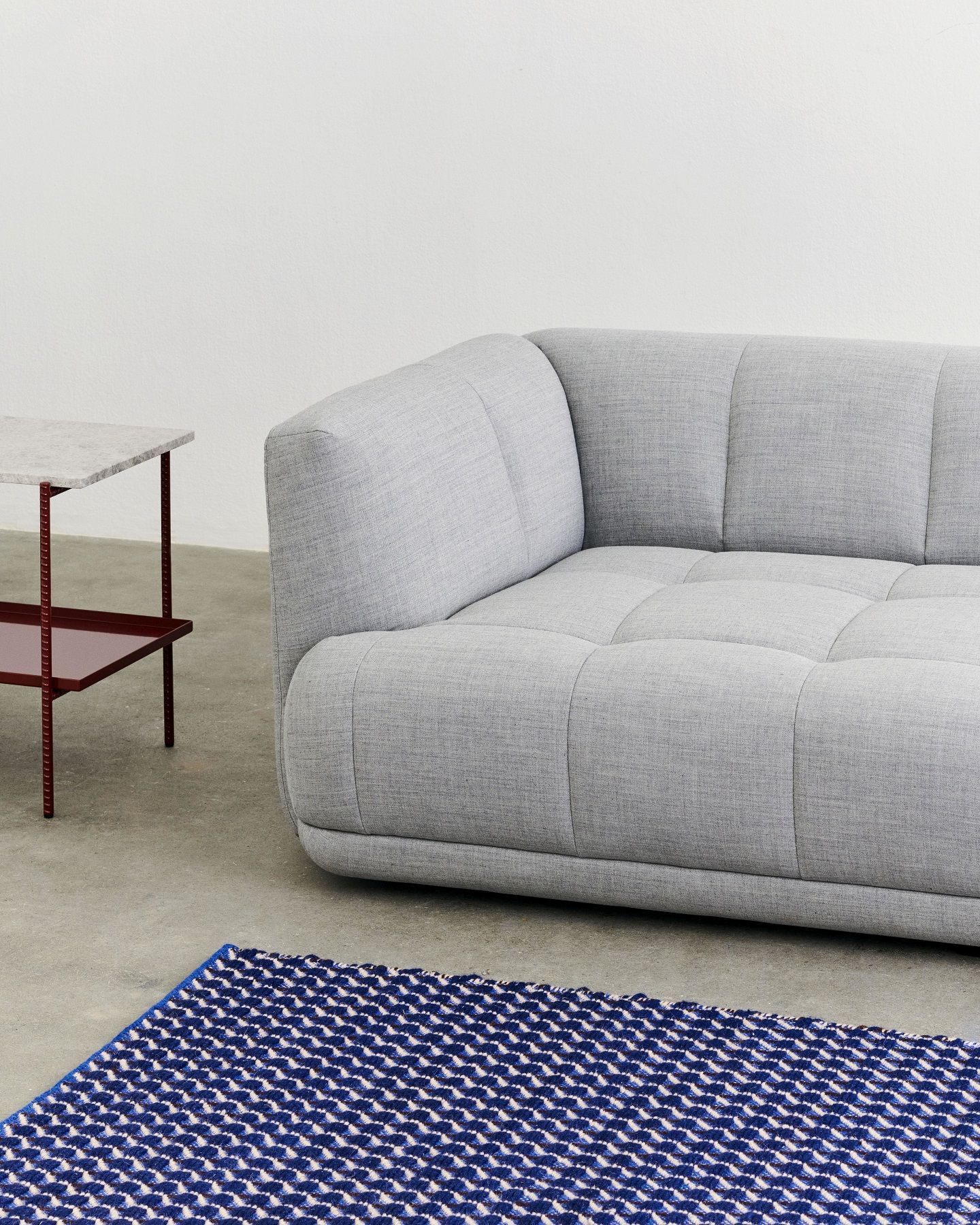 Nahaufnahme des Channel Rug Teppichs von Hay in Blau-Weiß mit geometrischem Muster, neben grauem Sofa.