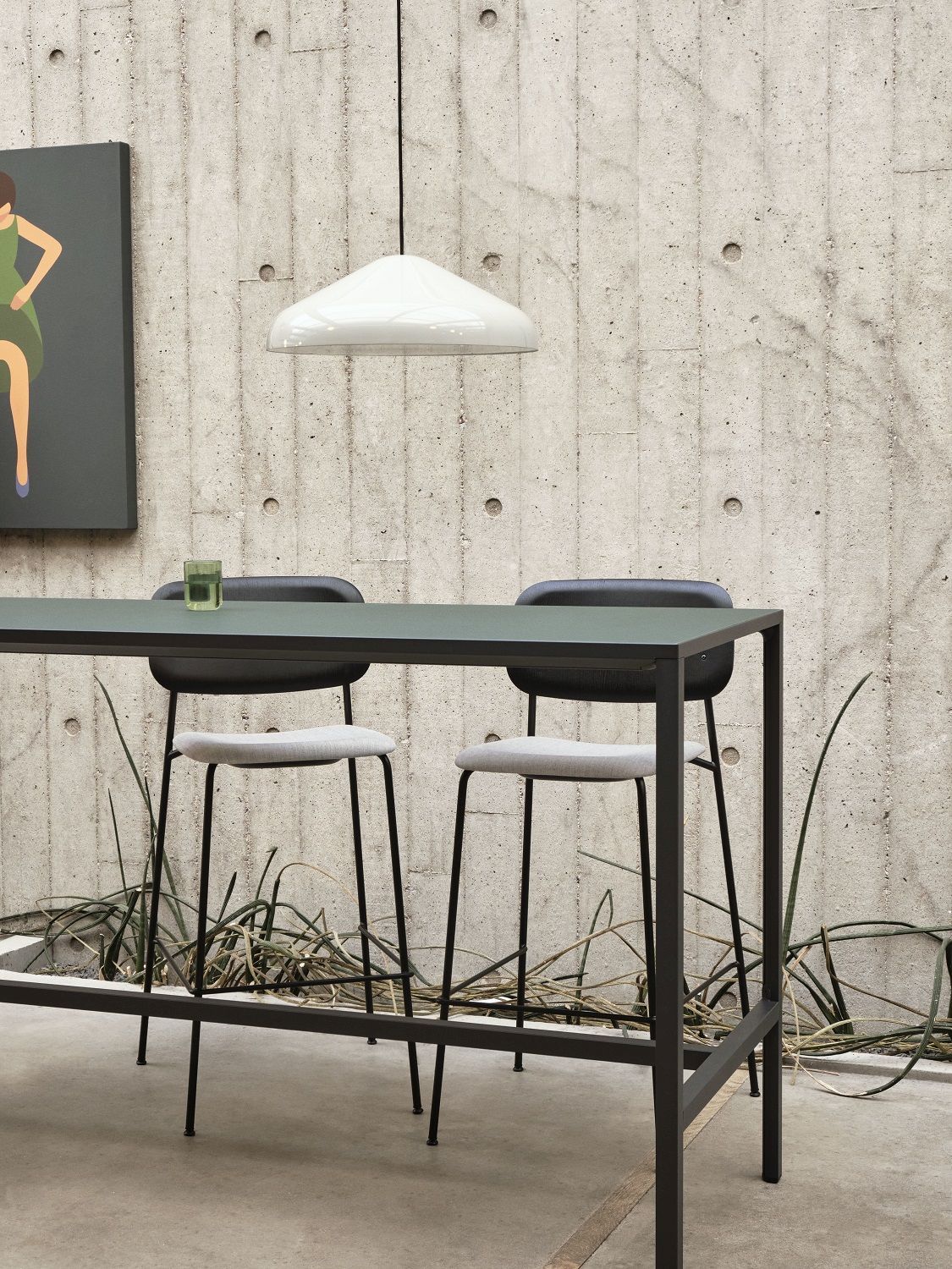 Zwei graue Hay Soft Edge Barhocker mit schwarzem Gestell an einem hohen Tisch vor Betonwand.