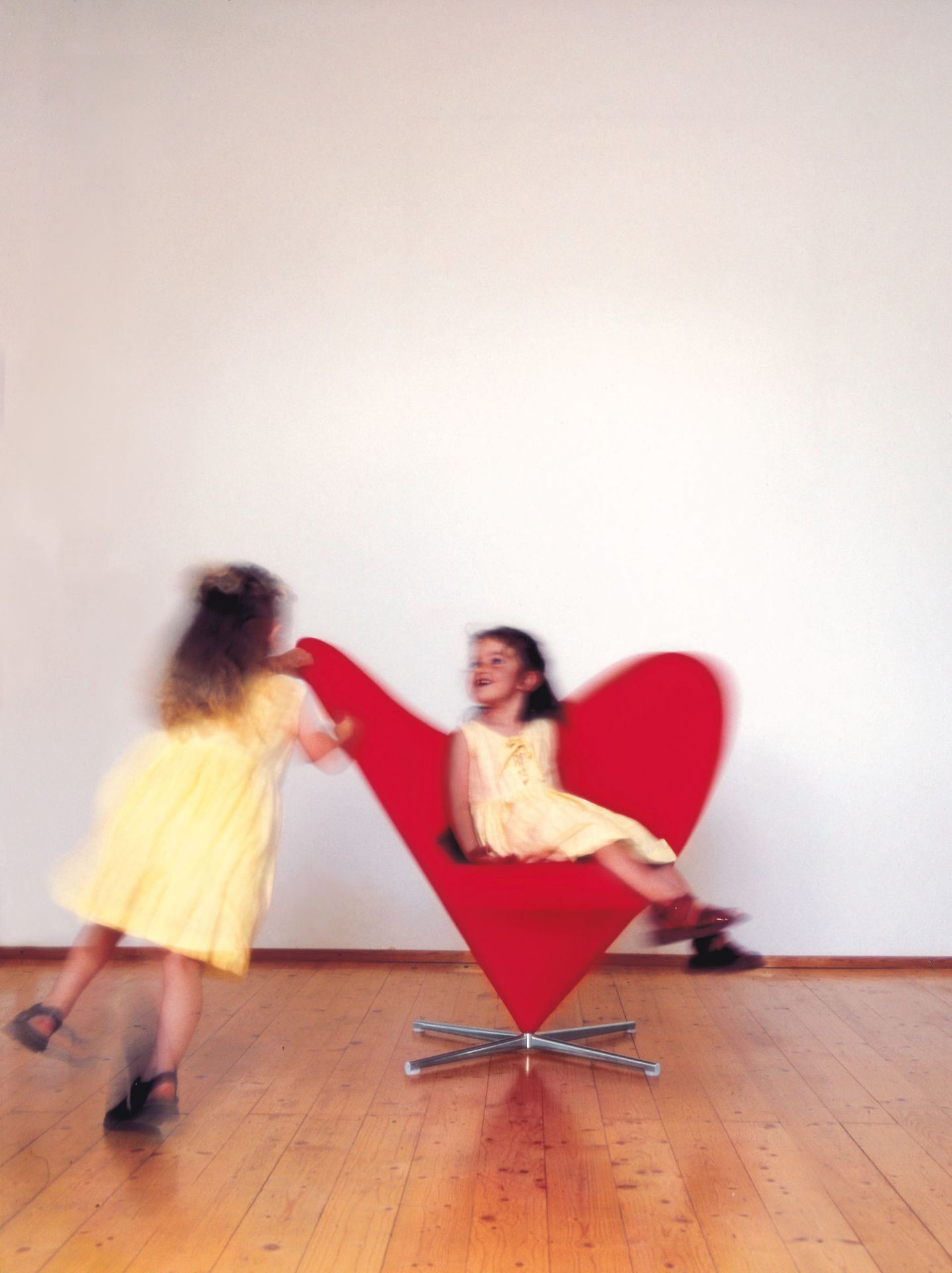 Zwei Mädchen spielen mit einem roten Heart Cone Chair von Vitra auf einem Holzboden.