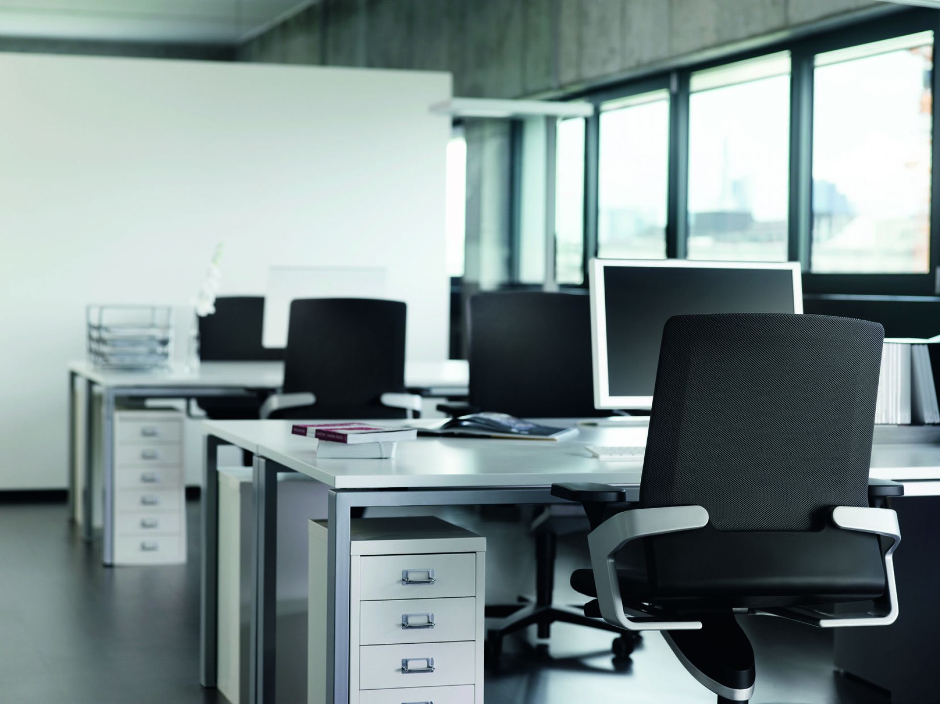 Modernes Büro mit ergonomischem Bürostuhl, weißen Schreibtischen, Rollcontainern und Computern für effizientes Arbeiten.