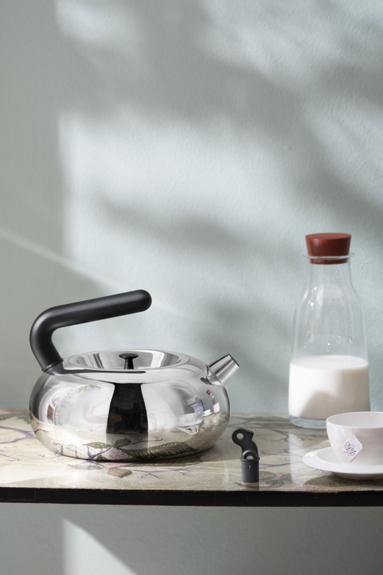 Alessi Irony Bulbul Wasserkessel aus Edelstahl, mit Milchflasche und Teetasse auf gemusterter Oberfläche.