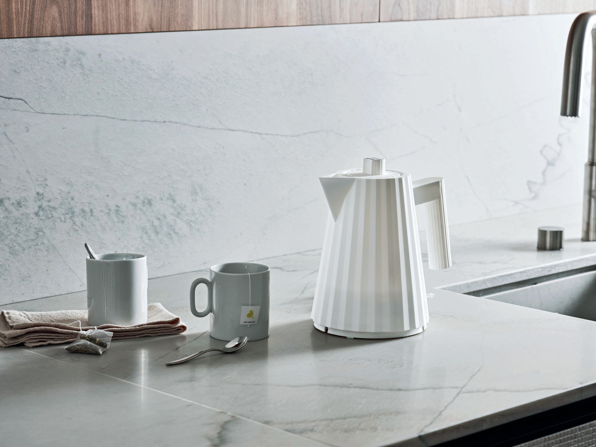 Eleganter, weißer Alessi Plissée Wasserkocher neben Tassen und Teebeuteln auf einer Marmor-Küchenarbeitsplatte.