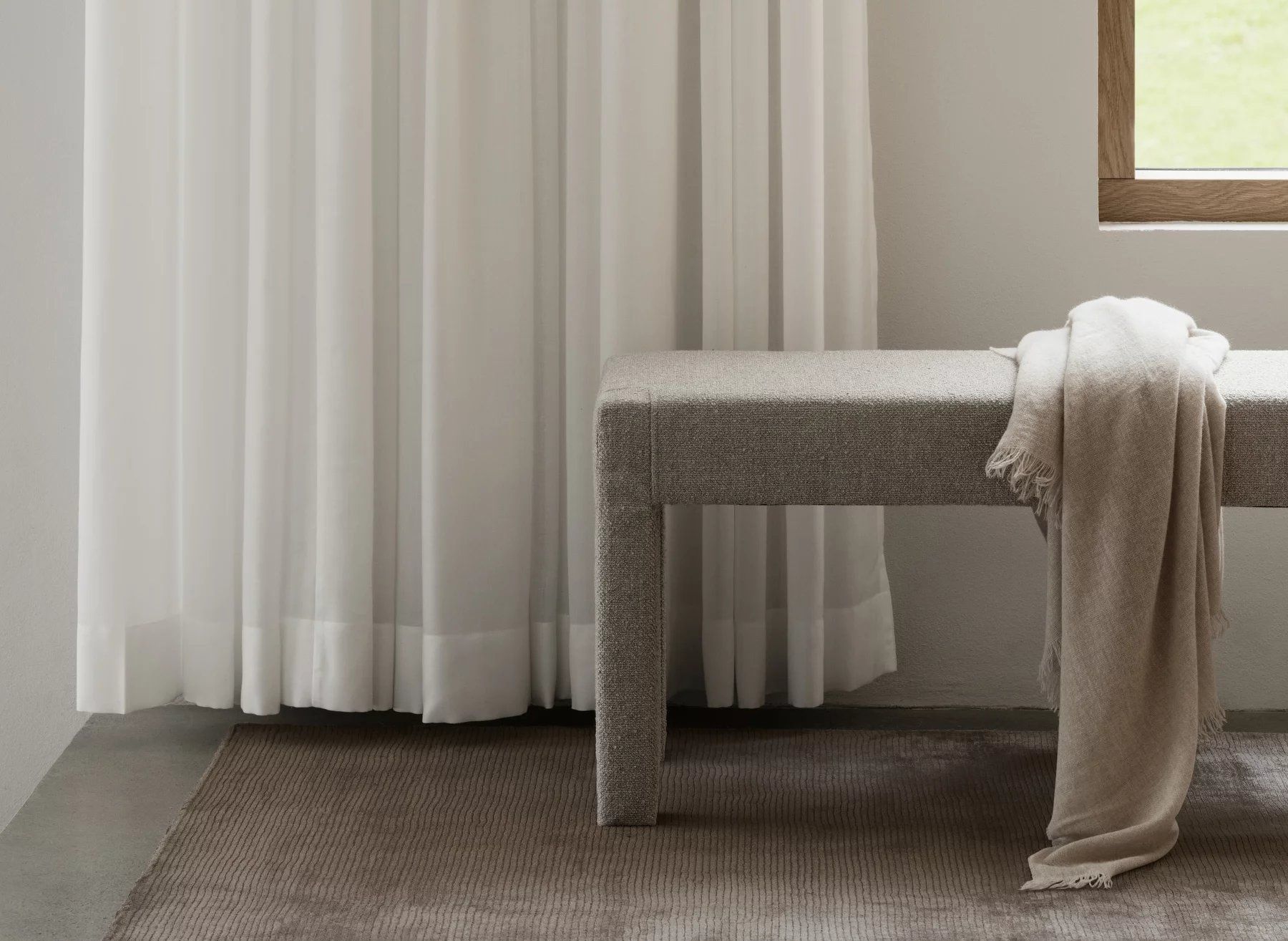 Blomus Sitta Bank mit beiger Decke in minimalistischem Interieur, helle Vorhänge und Teppich.