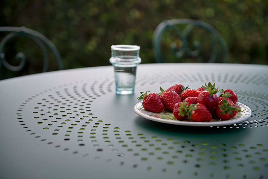 Nahaufnahme des Fermob 1900 Gartentisches mit Erdbeeren und einem Glas Wasser im Freien.